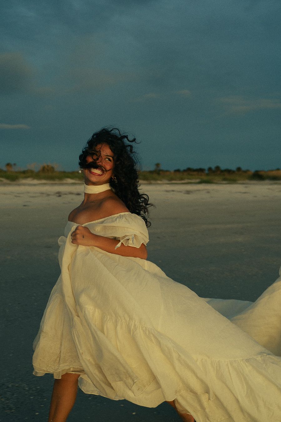 Woman in flowing white off-shoulder dress twirling on a beach at golden hour with dramatic cloudy sky.