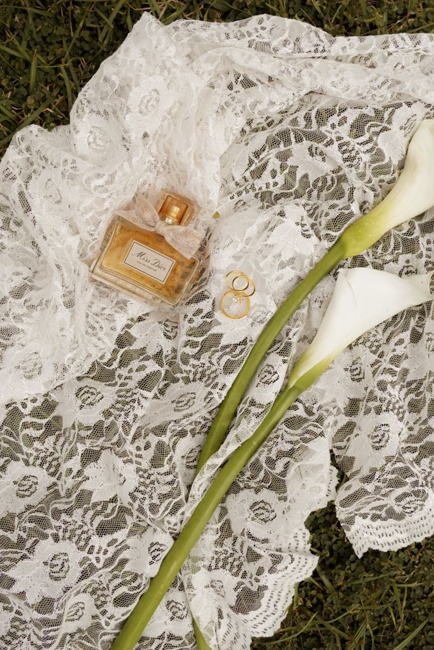 White lace wedding veil laid on grass with gold perfume bottle, wedding rings, and a white calla lily.