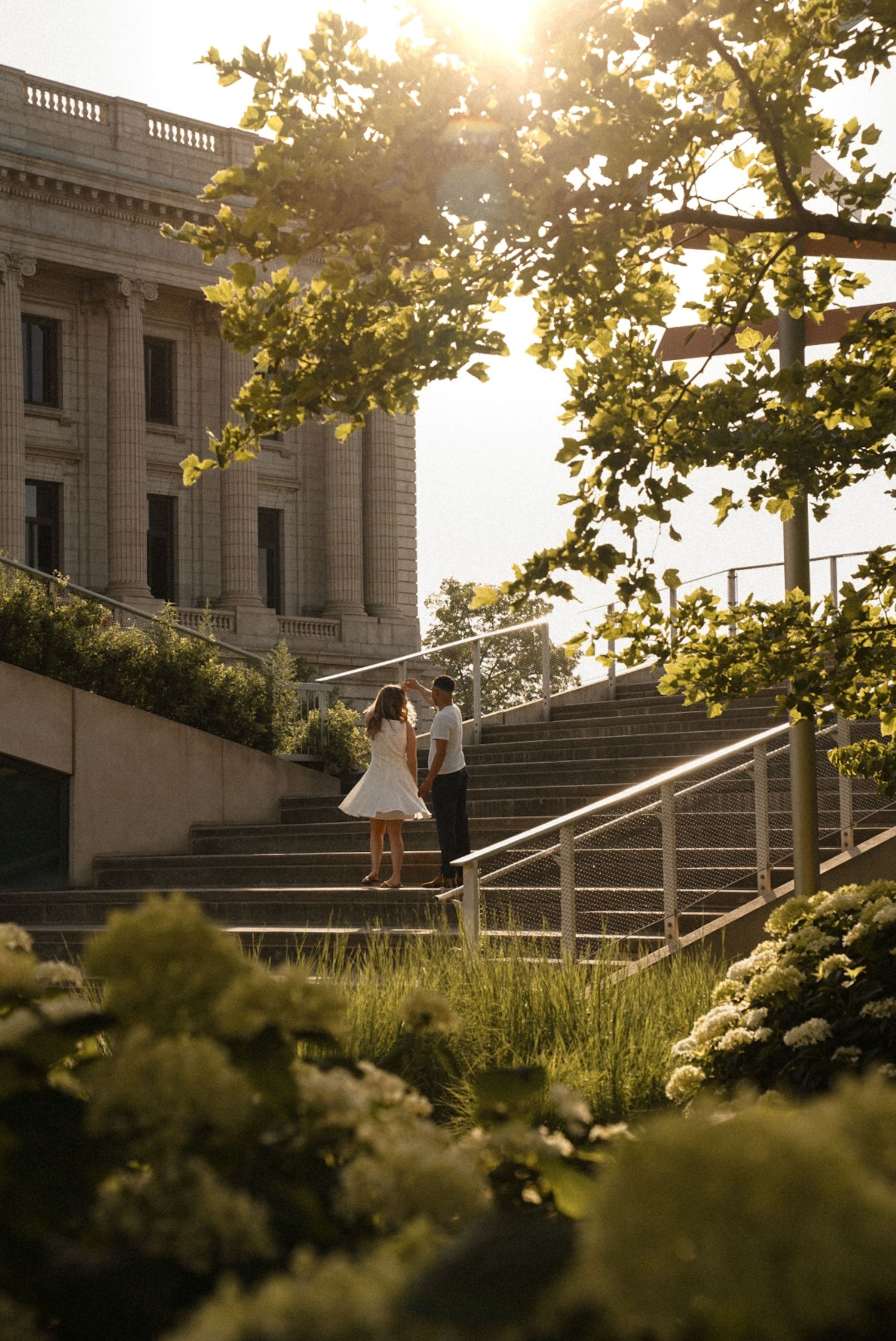 Couple sharing a romantic moment on sunlit stone steps beside a grand neoclassical building surrounded by lush greenery.