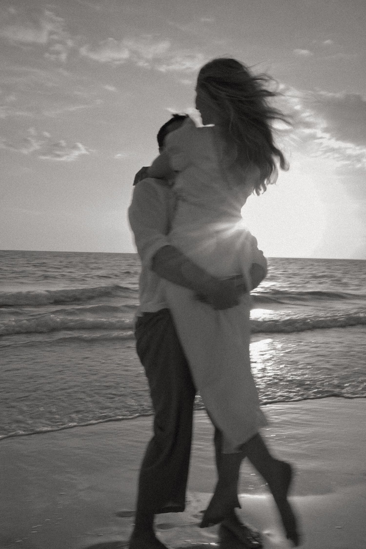Black and white photo of a romantic couple embracing on a beach at sunset with ocean waves in the background.