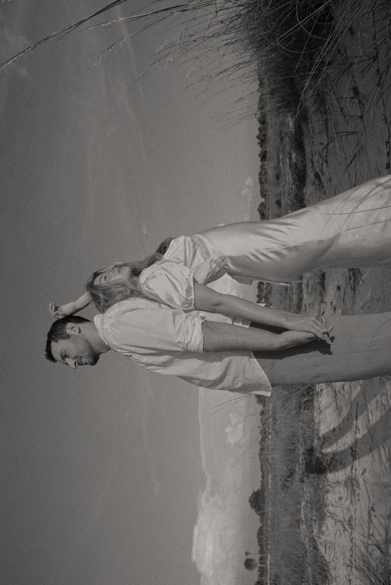 Black and white photo of a couple embracing on a sandy beach, dressed in light summer clothing.
