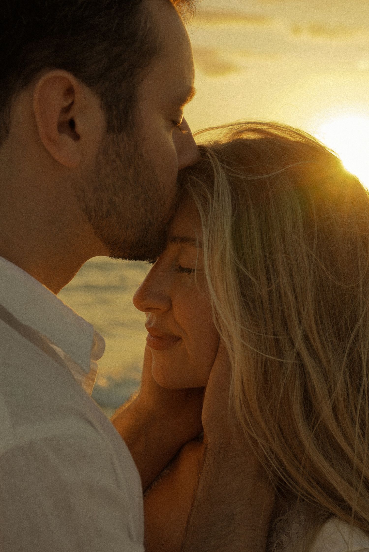 Romantic couple sharing a tender moment at golden sunset, man kissing woman's forehead on a beach.
