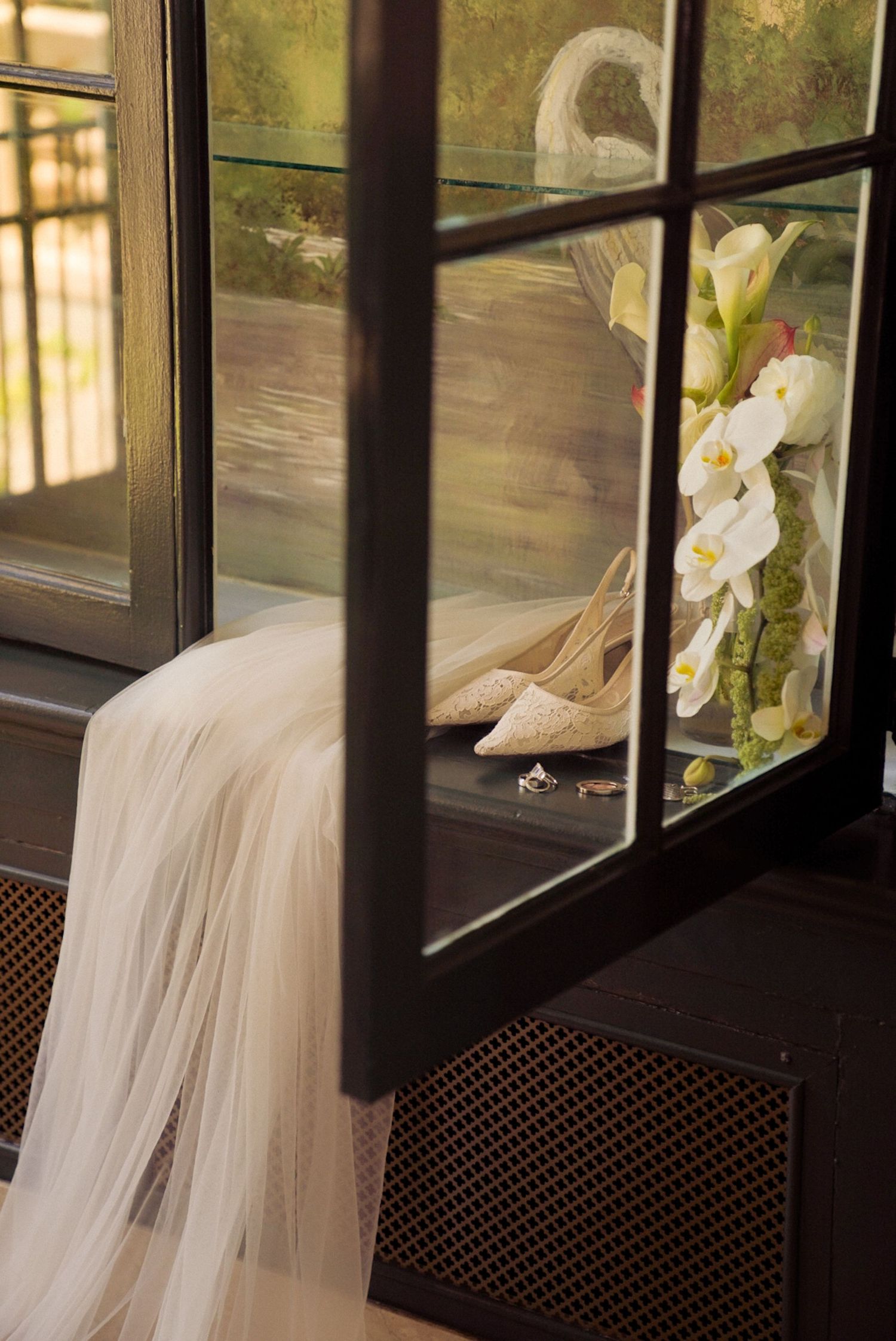 Bridal veil draped over chair beside glass window with white heels, wedding rings, and orchid bouquet arrangement.