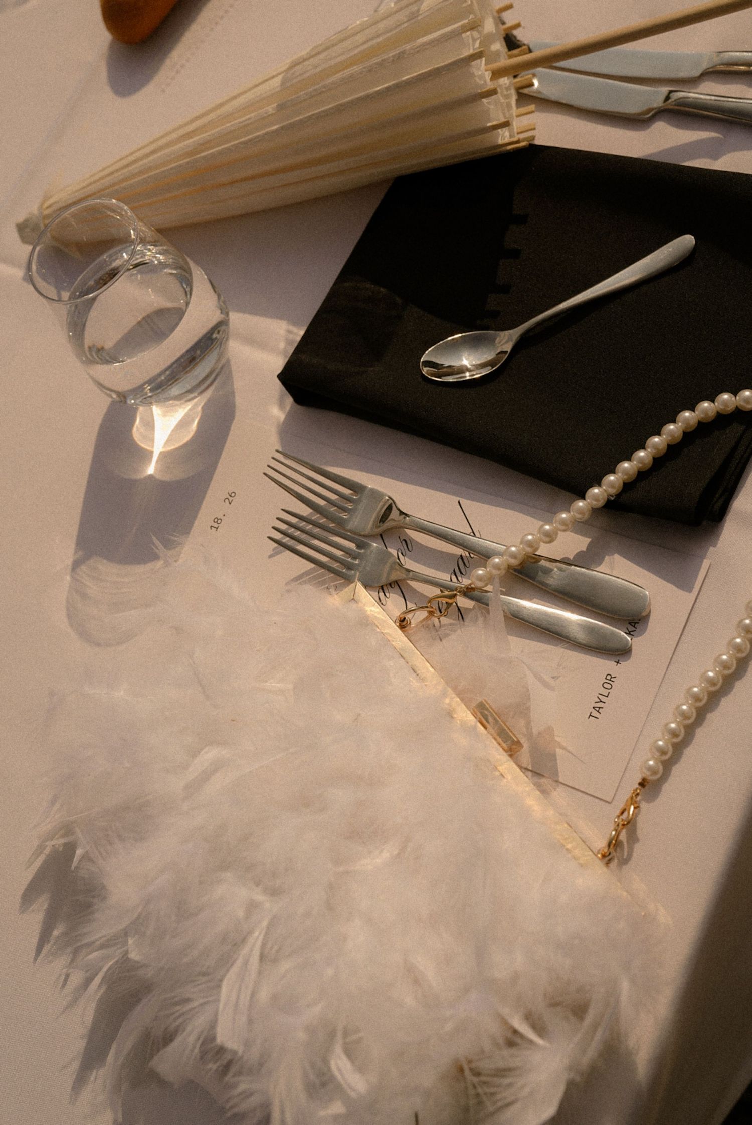 Elegant table setting with white feathers, pearl necklace, silver cutlery, black napkin, and decorative umbrella.