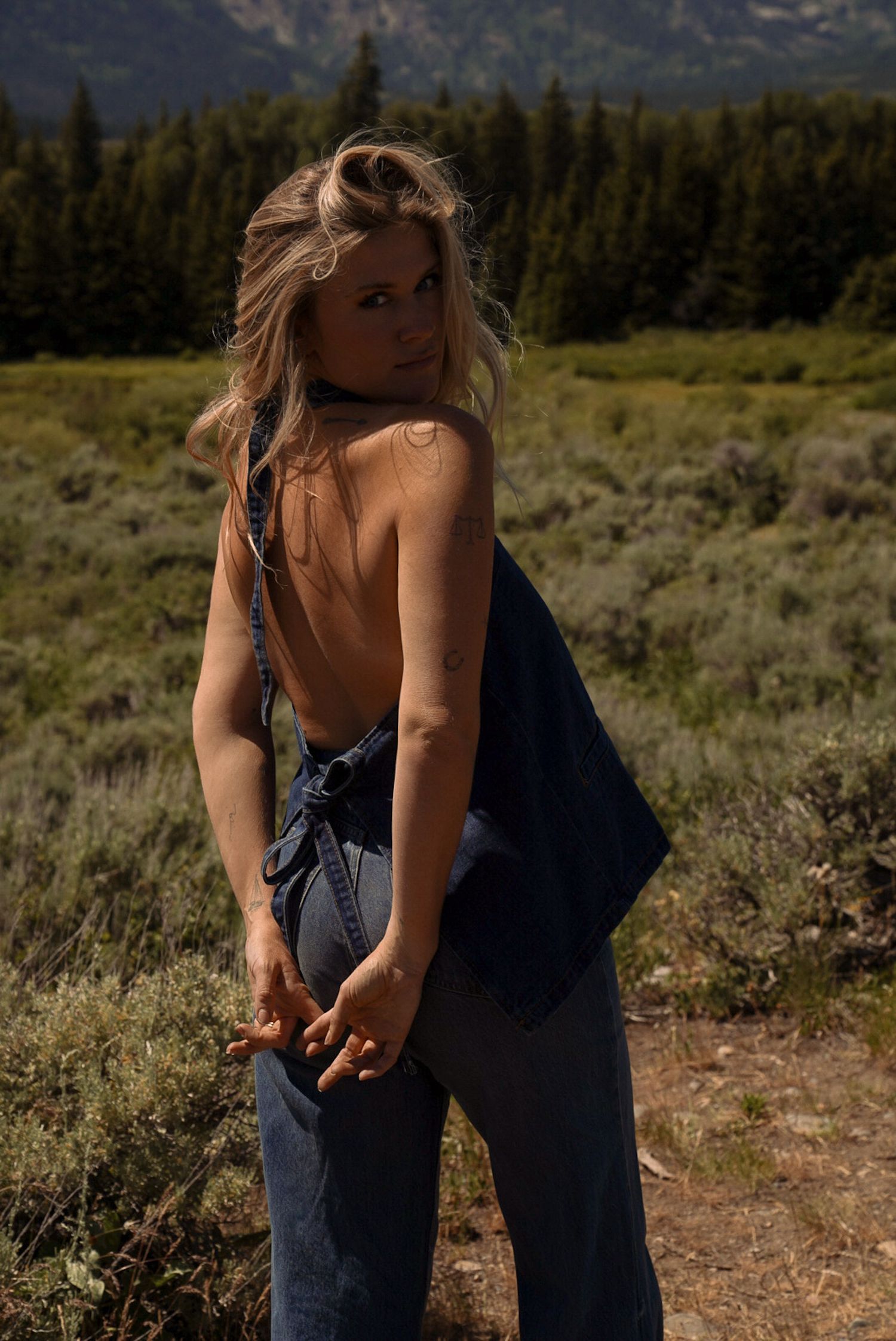 Woman in backless navy top and jeans poses in mountain meadow with pine trees backdrop.