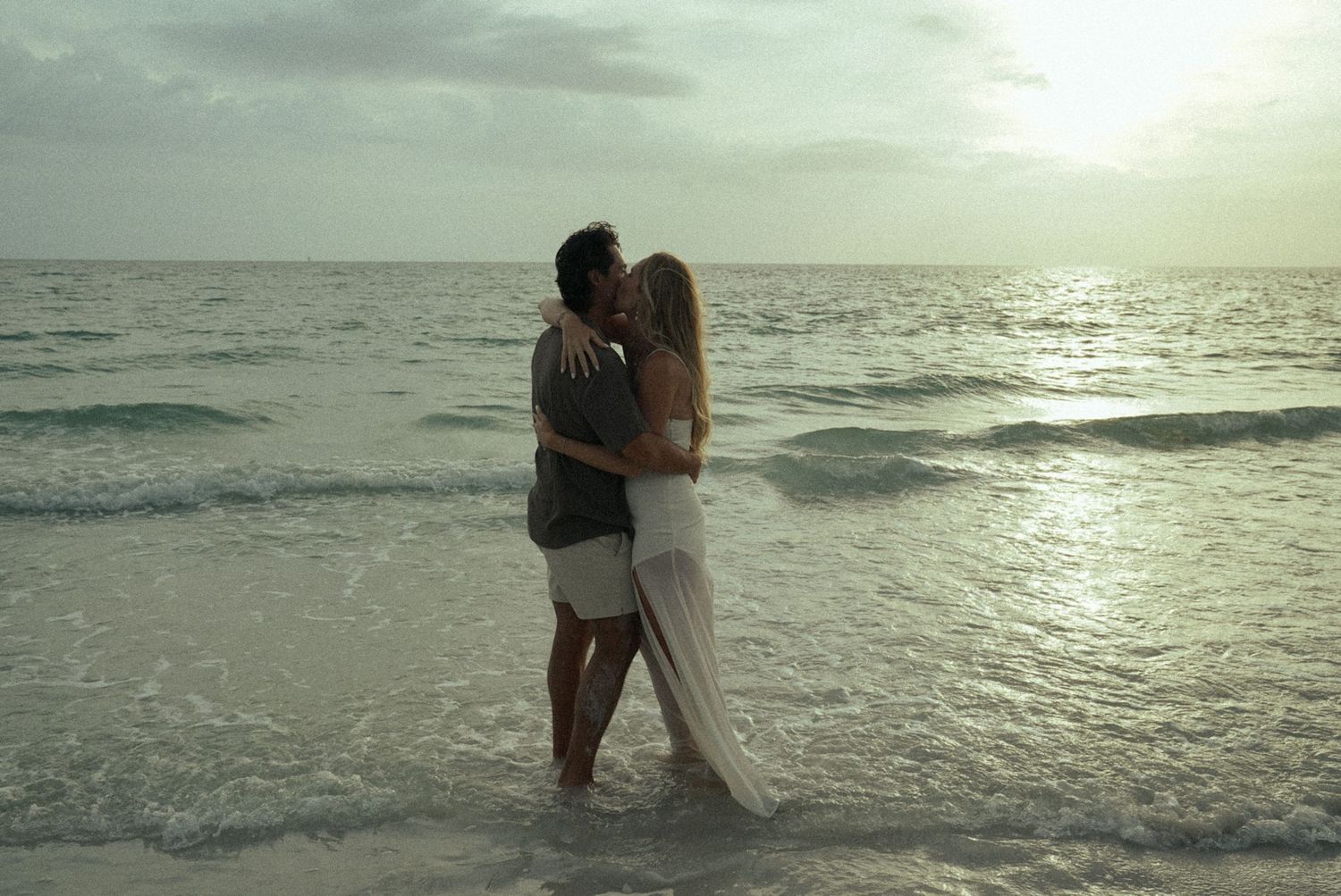 Romantic couple kissing on a tropical beach at sunset, standing in shallow ocean waves.