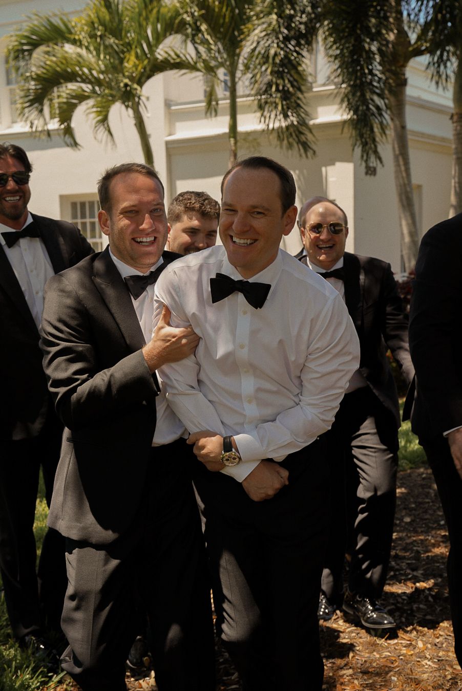 Groom in white shirt and bow tie laughing with groomsmen in black tuxedos outdoors at a wedding.