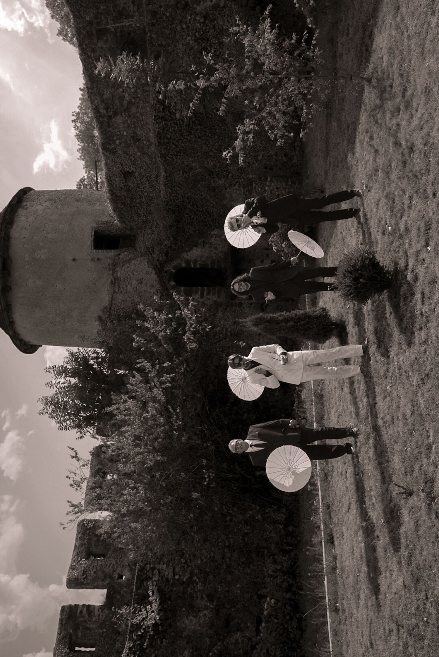 Black and white photo of people in vintage attire holding parasols near a historic stone tower outdoors.