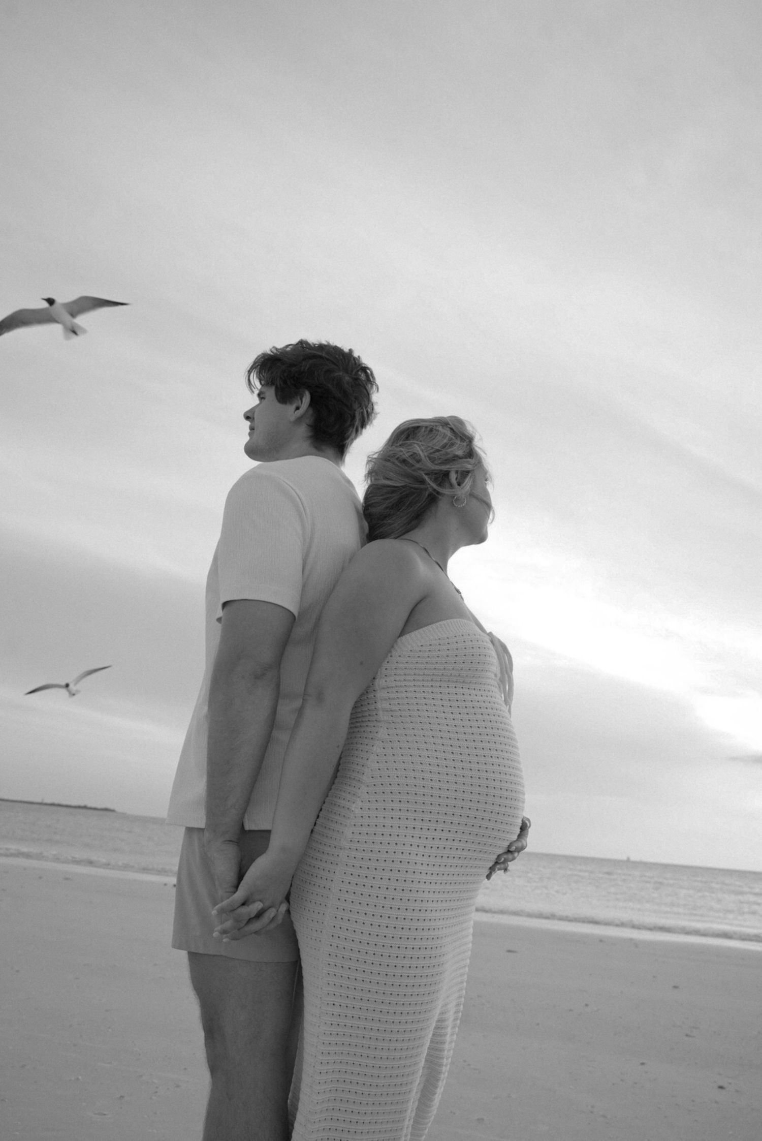 Pregnant couple holding hands on a beach at sunset with seagulls flying overhead in black and white.
