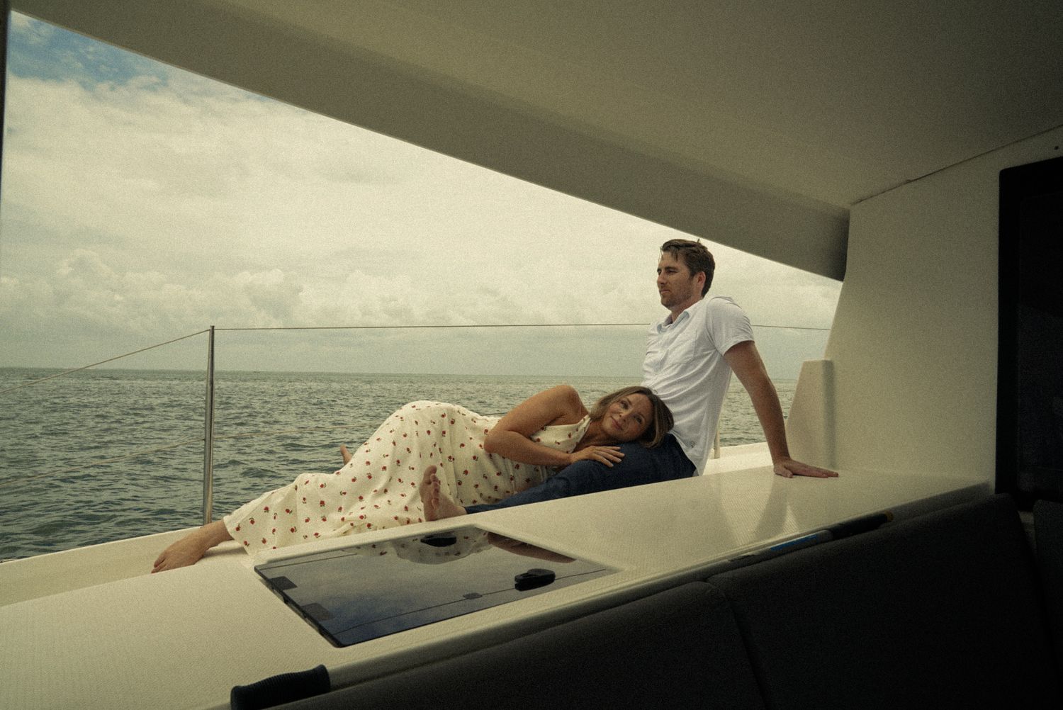 Couple relaxing on a luxury yacht deck overlooking calm ocean waters, woman in floral dress reclining beside seated man.