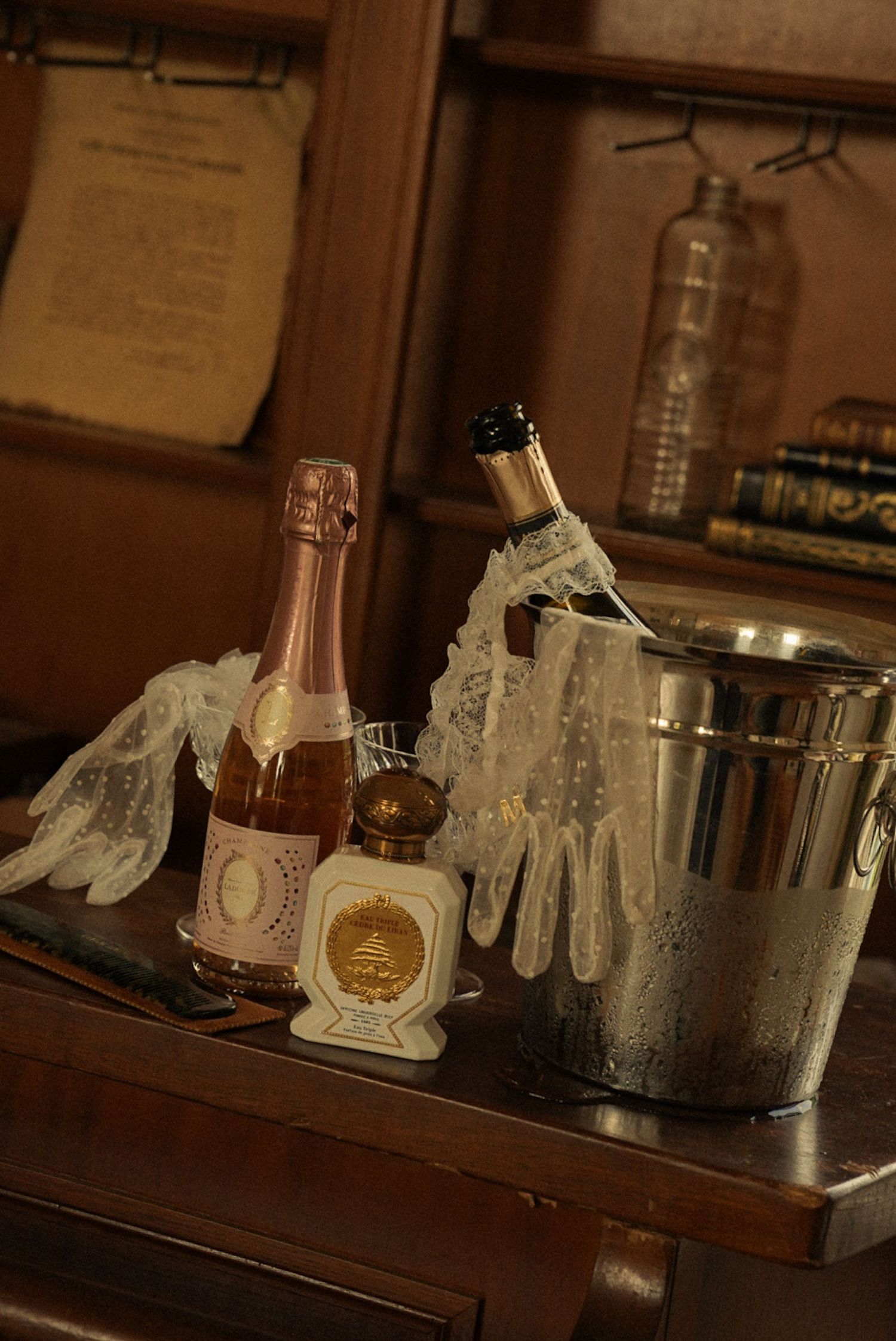 Vintage bar setup with rosé champagne, ice bucket, lace gloves, and perfume bottle on wooden dresser.