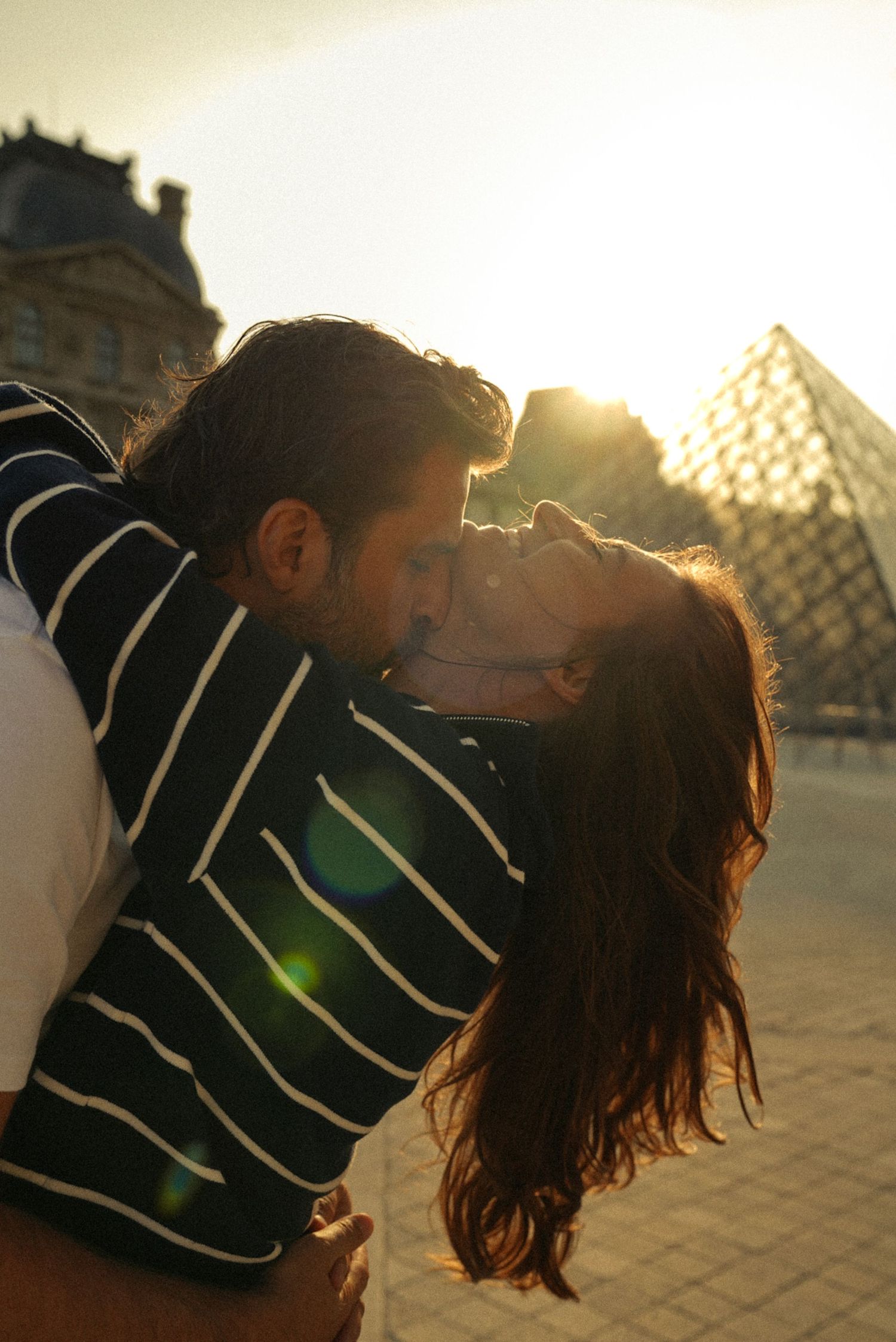 Couple sharing a romantic kiss at sunset near the Louvre pyramid in Paris, with golden lens flare.