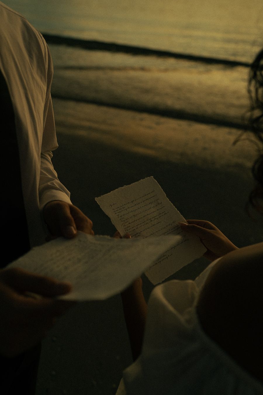 Two people reading handwritten letters together on a beach at golden sunset hour.