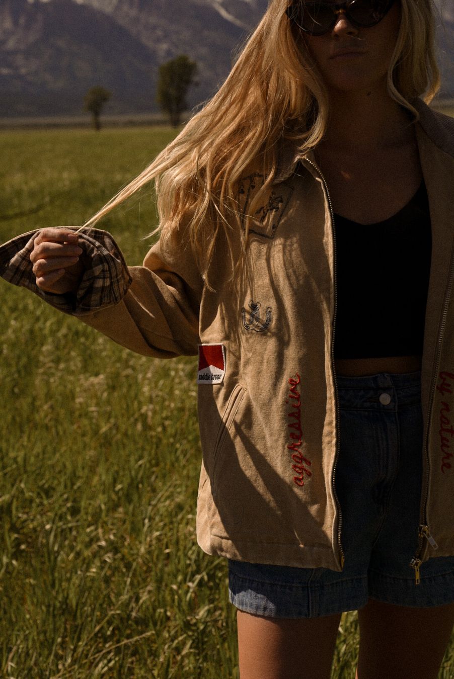 Blonde woman in tan jacket and denim shorts standing in a green field with mountains in the background.