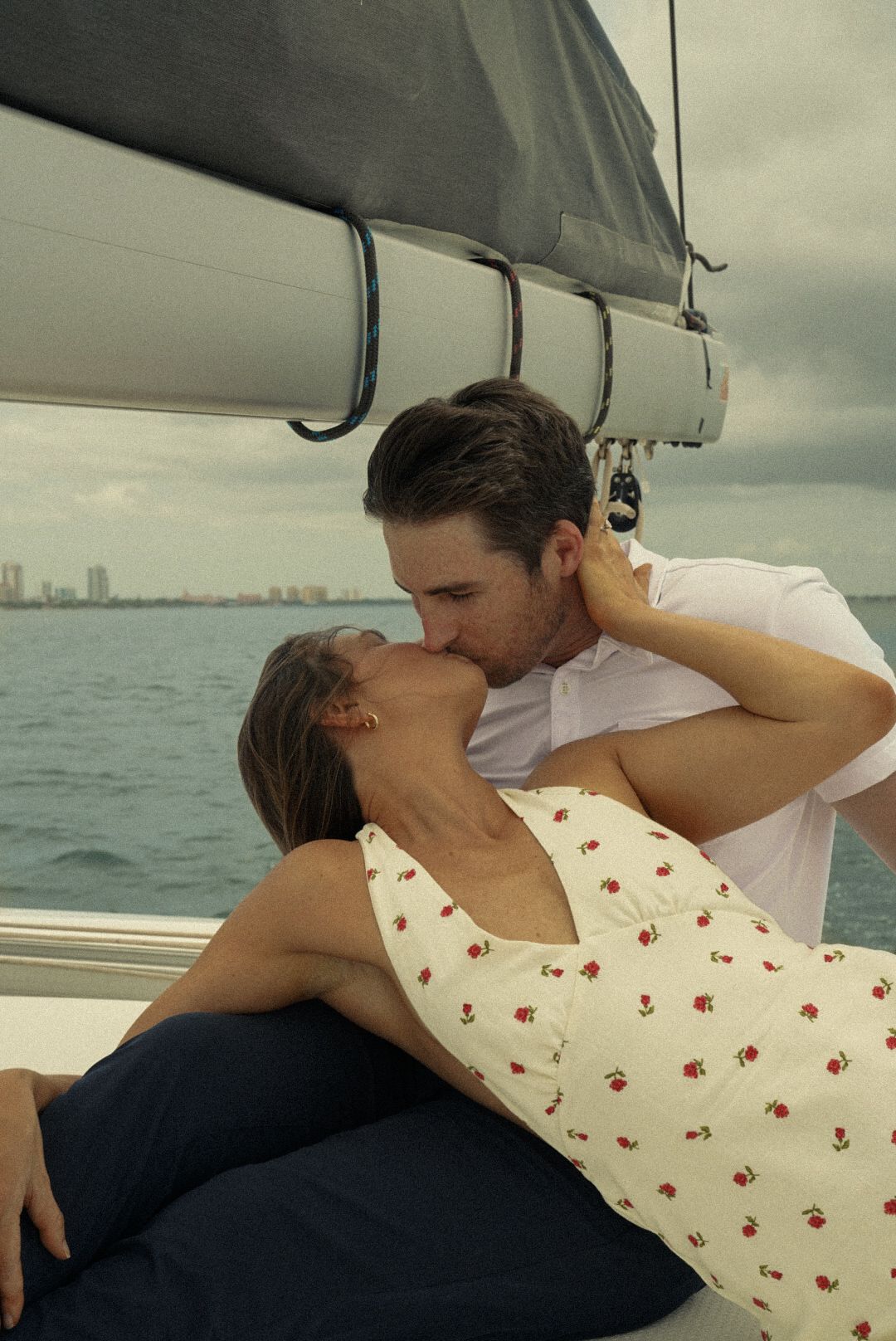 Romantic couple sharing a kiss on a sailboat deck with ocean and city skyline in the background.