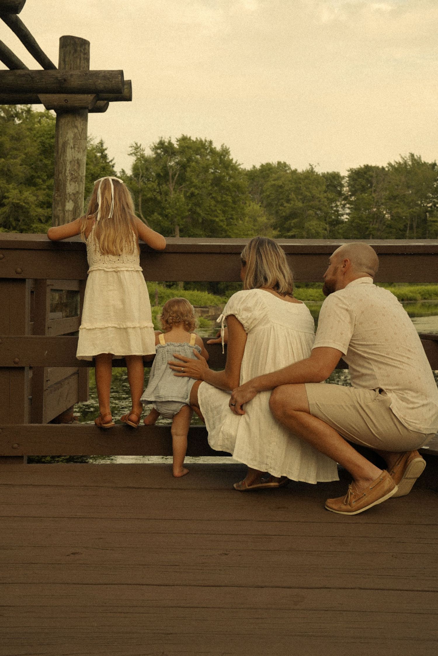 Family of four on a wooden bridge deck overlooking a lush green pond, parents watching two young girls lean on the railing.