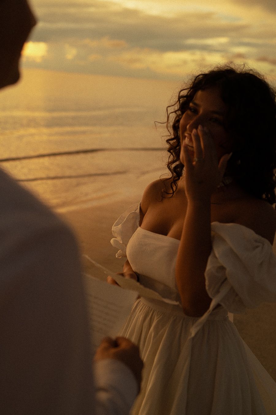 Couple sharing romantic moment on beach at golden sunset, woman in white off-shoulder dress smiling emotionally.