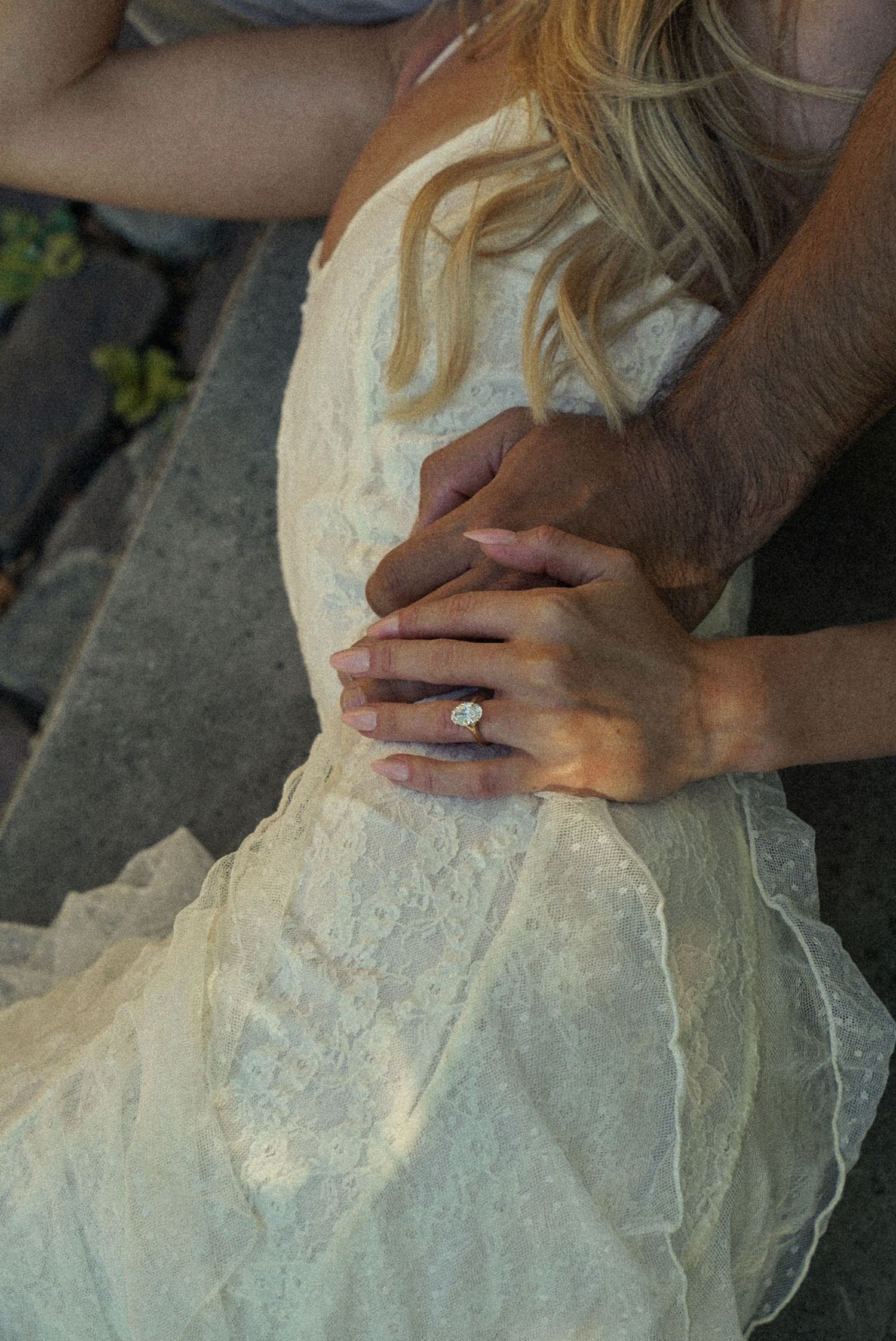 Close-up of bride in lace wedding dress holding hands with groom, showcasing a large diamond engagement ring.