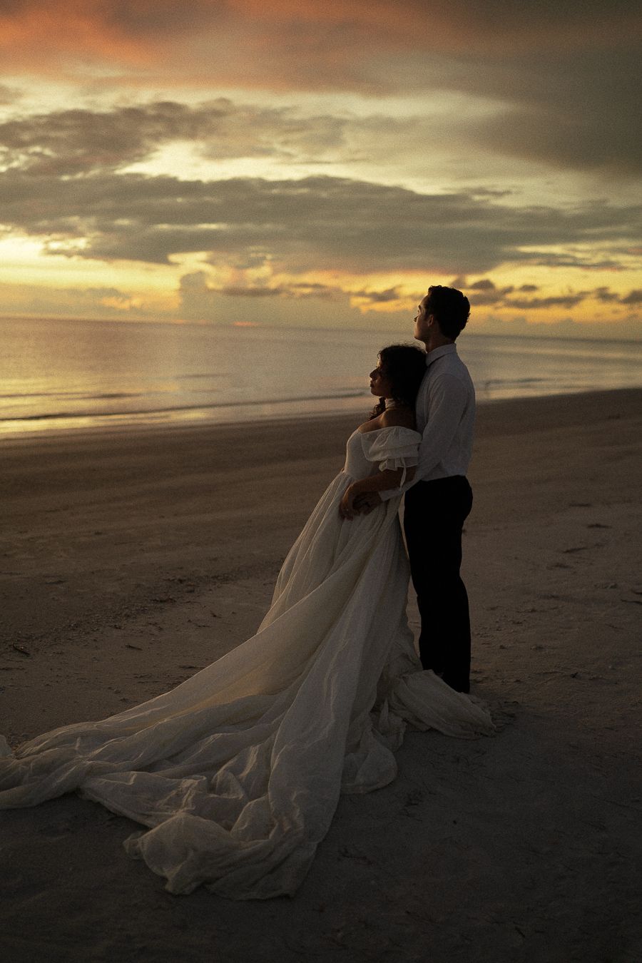 A bride and groom embrace on a sandy beach at sunset, her white gown trailing behind them under a dramatic cloudy sky.