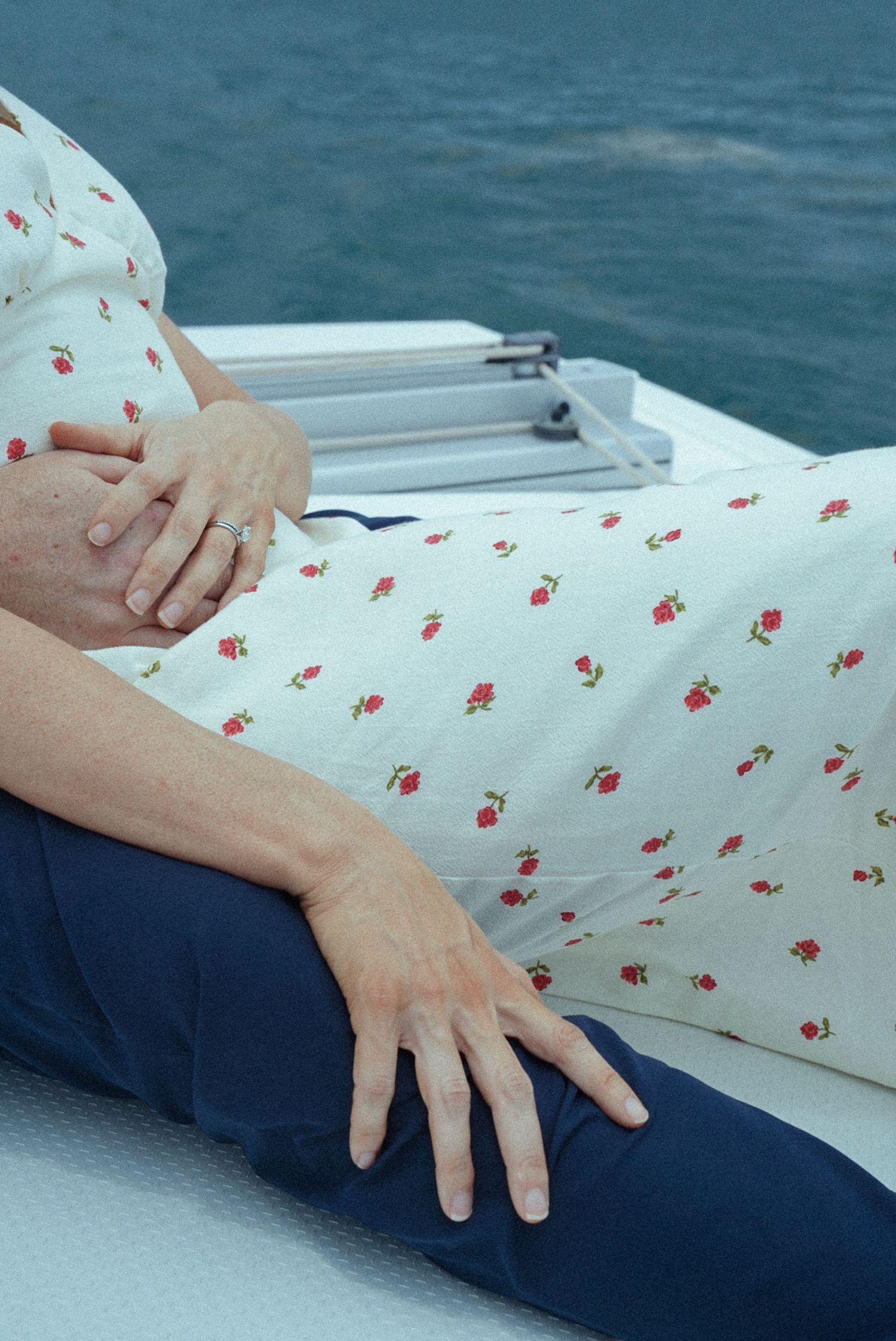 Pregnant woman in floral dress sitting on boat deck, hands resting on baby bump with ocean in background.