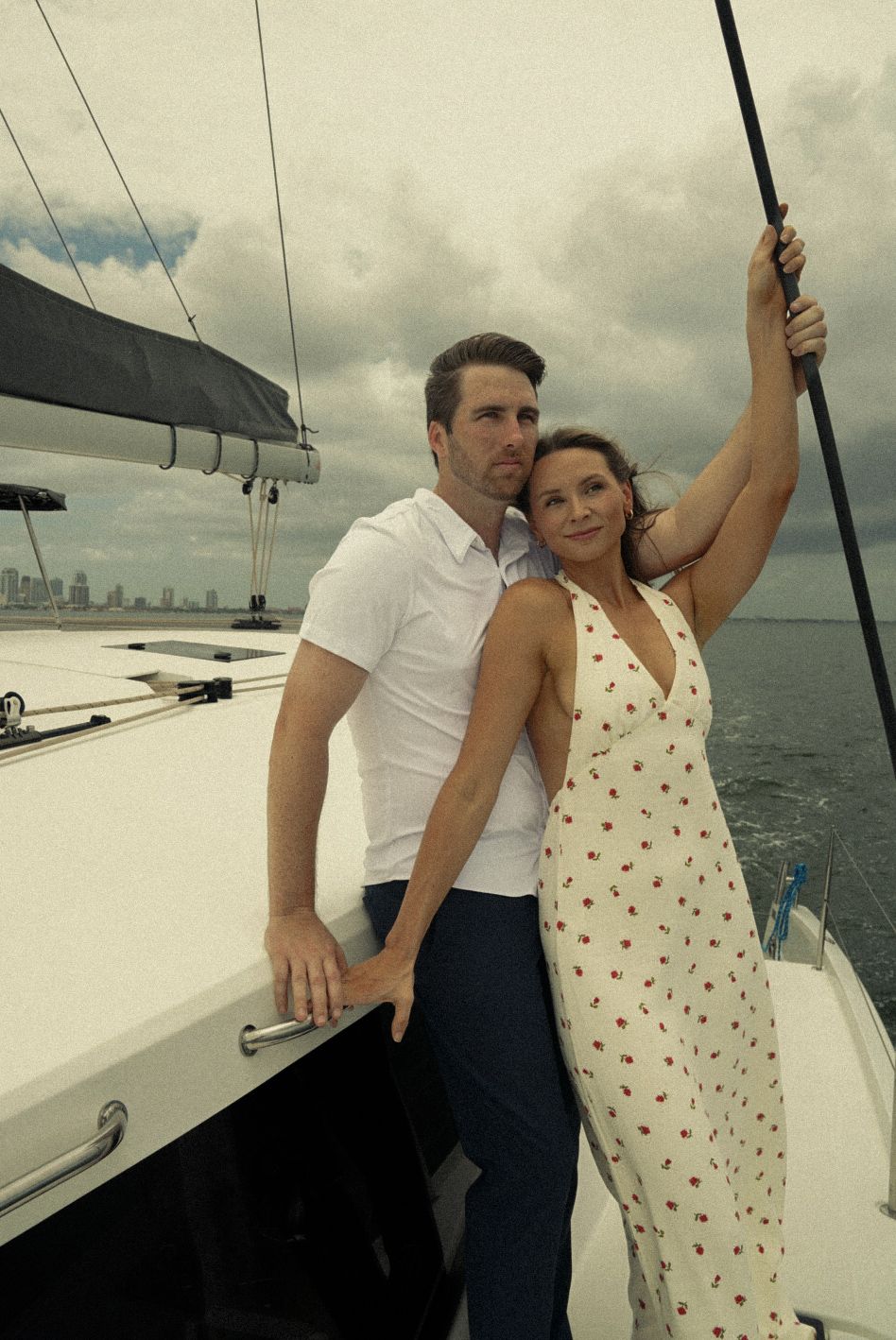 Couple posing romantically on a sailboat deck with ocean and city skyline in the background on a cloudy day.