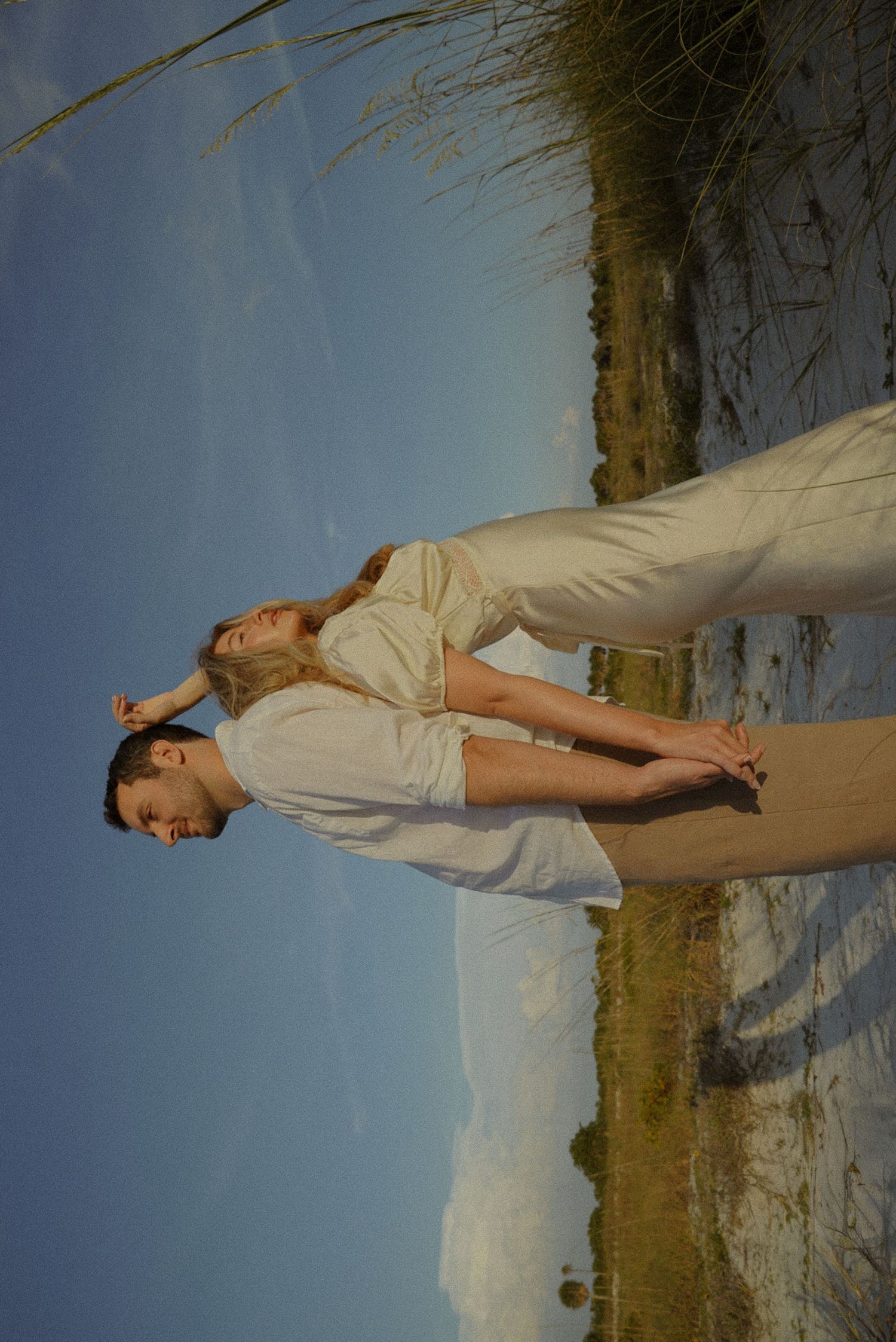 A couple in white attire poses romantically on a sandy beach with blue sky and coastal grass backdrop.