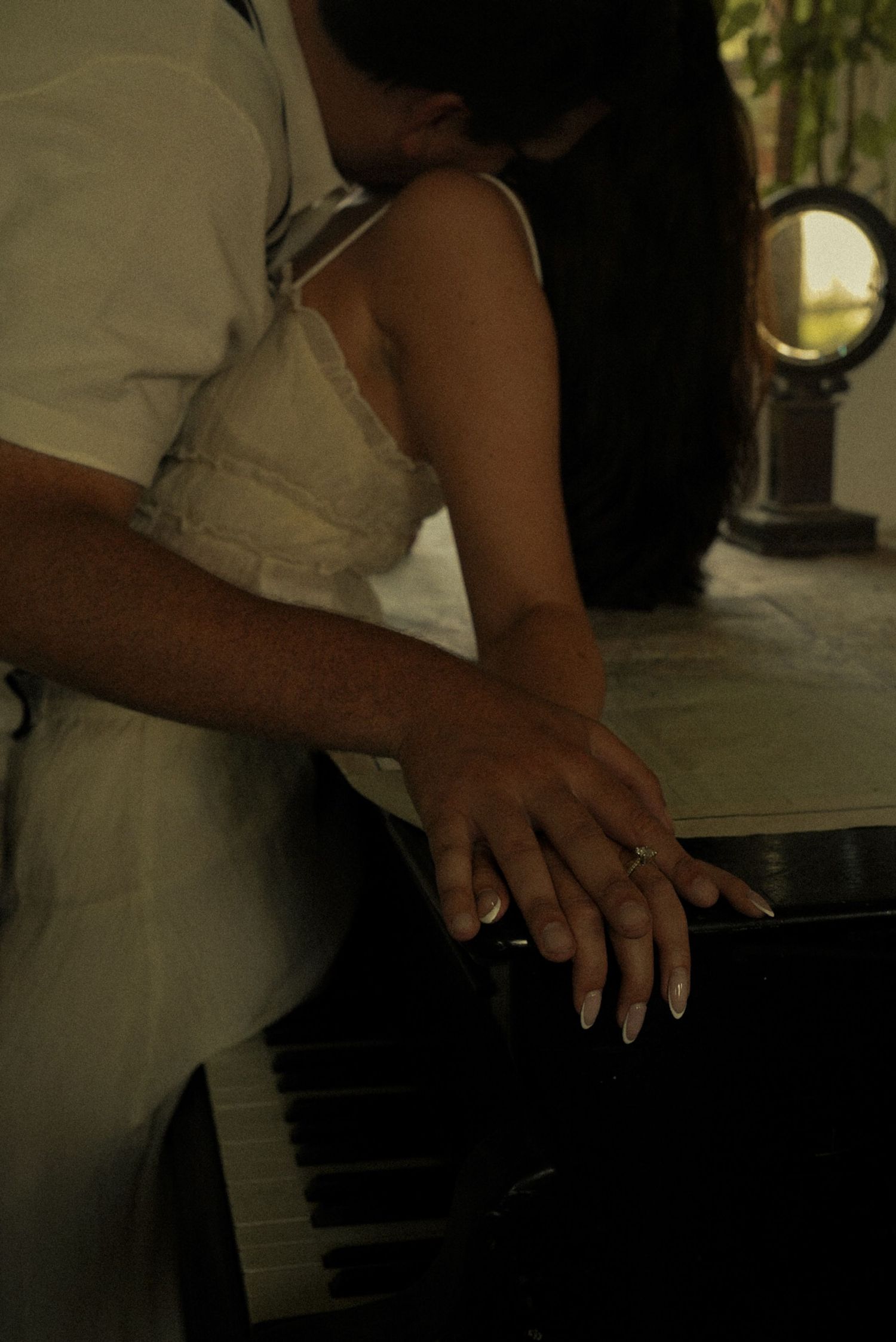 A couple's hands rest together on a black piano keys, woman wearing a ring, in a softly lit romantic setting.