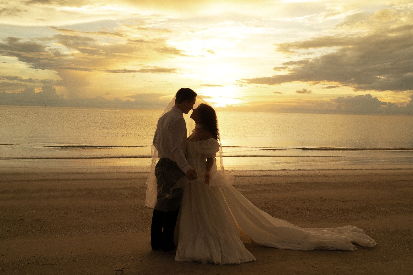 Bride and groom silhouetted against a golden sunset on a beach, sharing a romantic moment in wedding attire.