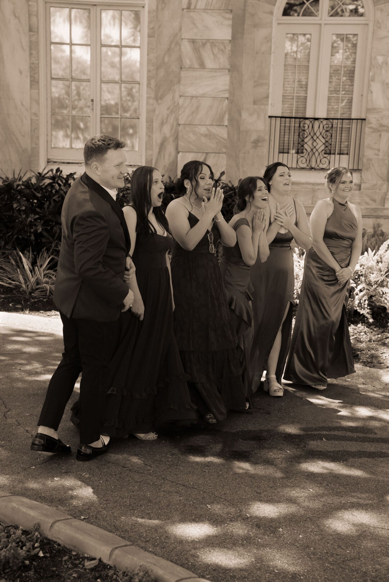 Wedding party in formal attire laughing outdoors near elegant stone building in sepia tone.