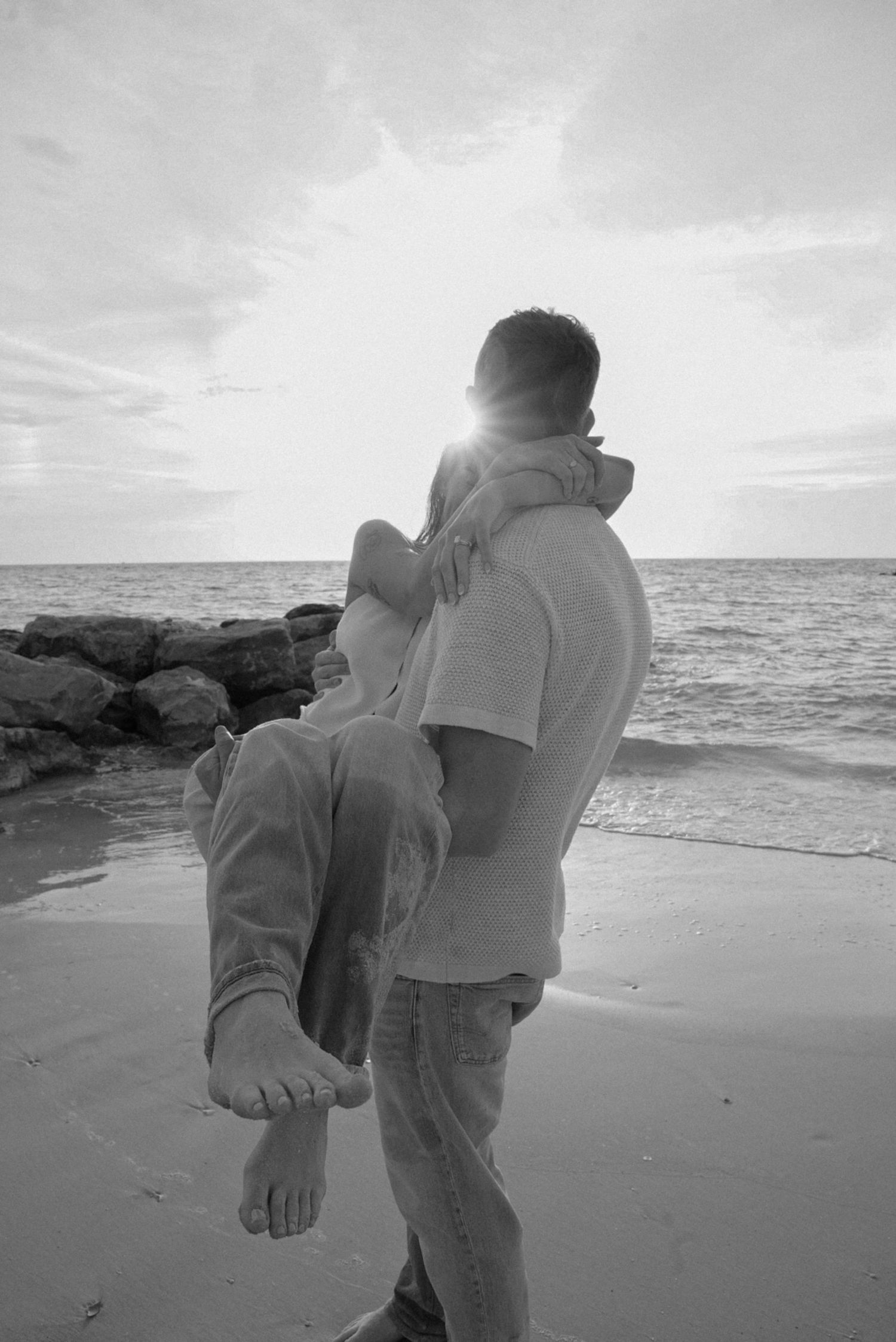 Black and white photo of a man carrying a child on a sandy beach at sunset with ocean waves in the background.