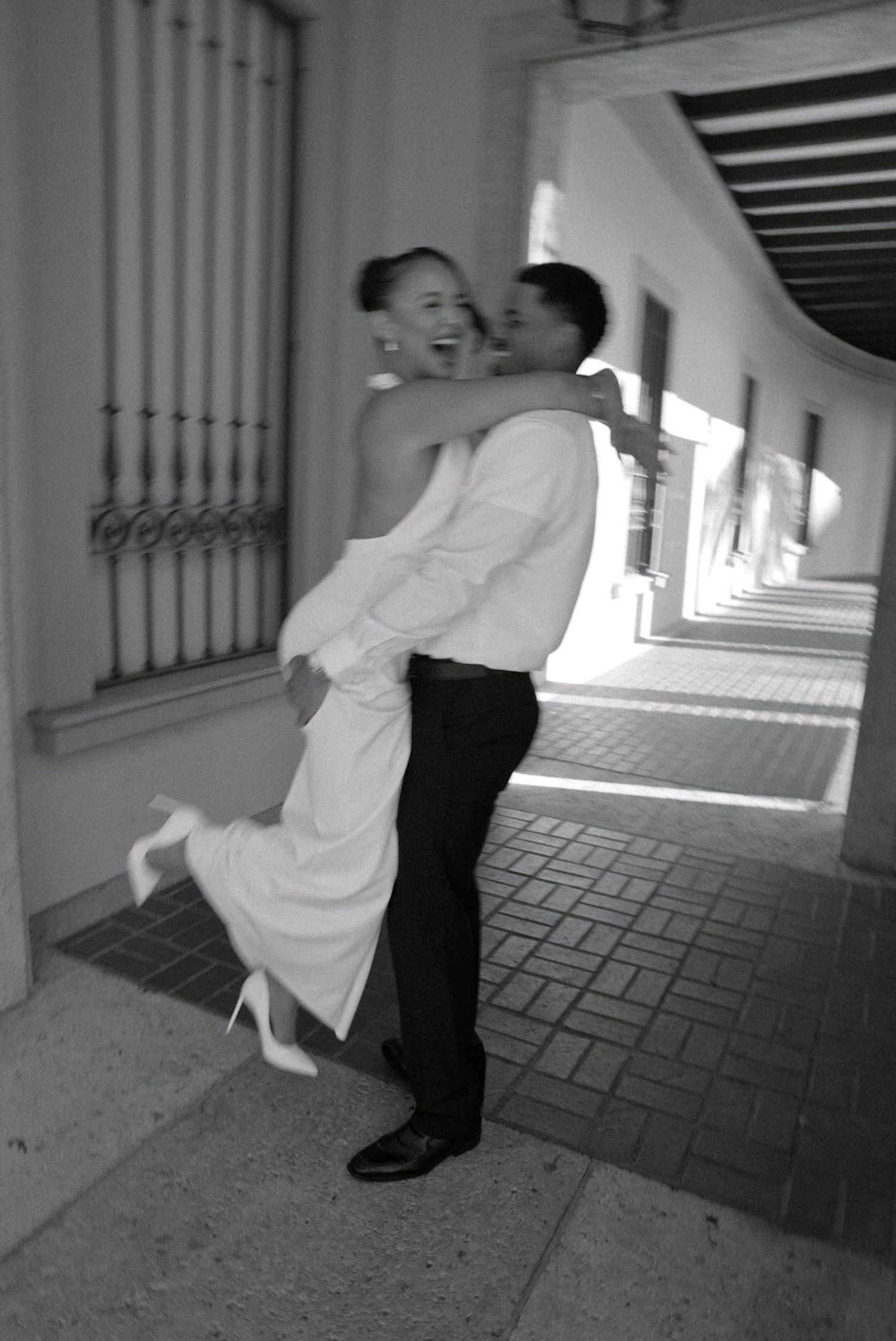Black and white photo of a groom lifting his laughing bride in a white dress along a sunlit archway corridor.