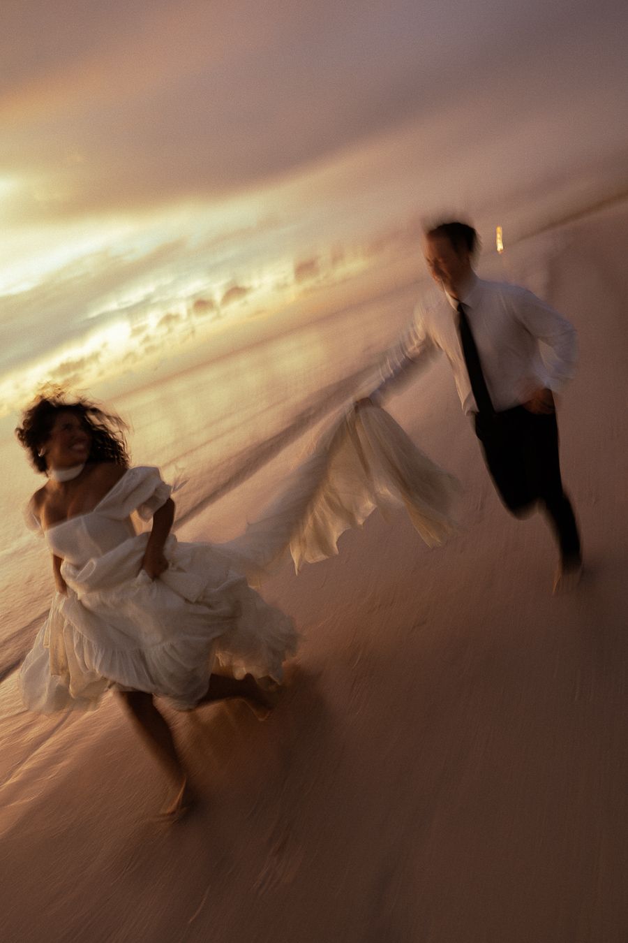Bride and groom running joyfully on a sandy beach at sunset, wedding dress flowing in the wind.