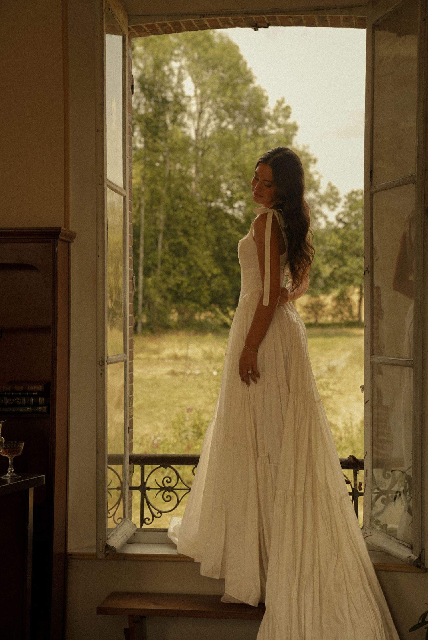 Bride in elegant white flowing gown standing in open French doors overlooking lush garden at golden hour.