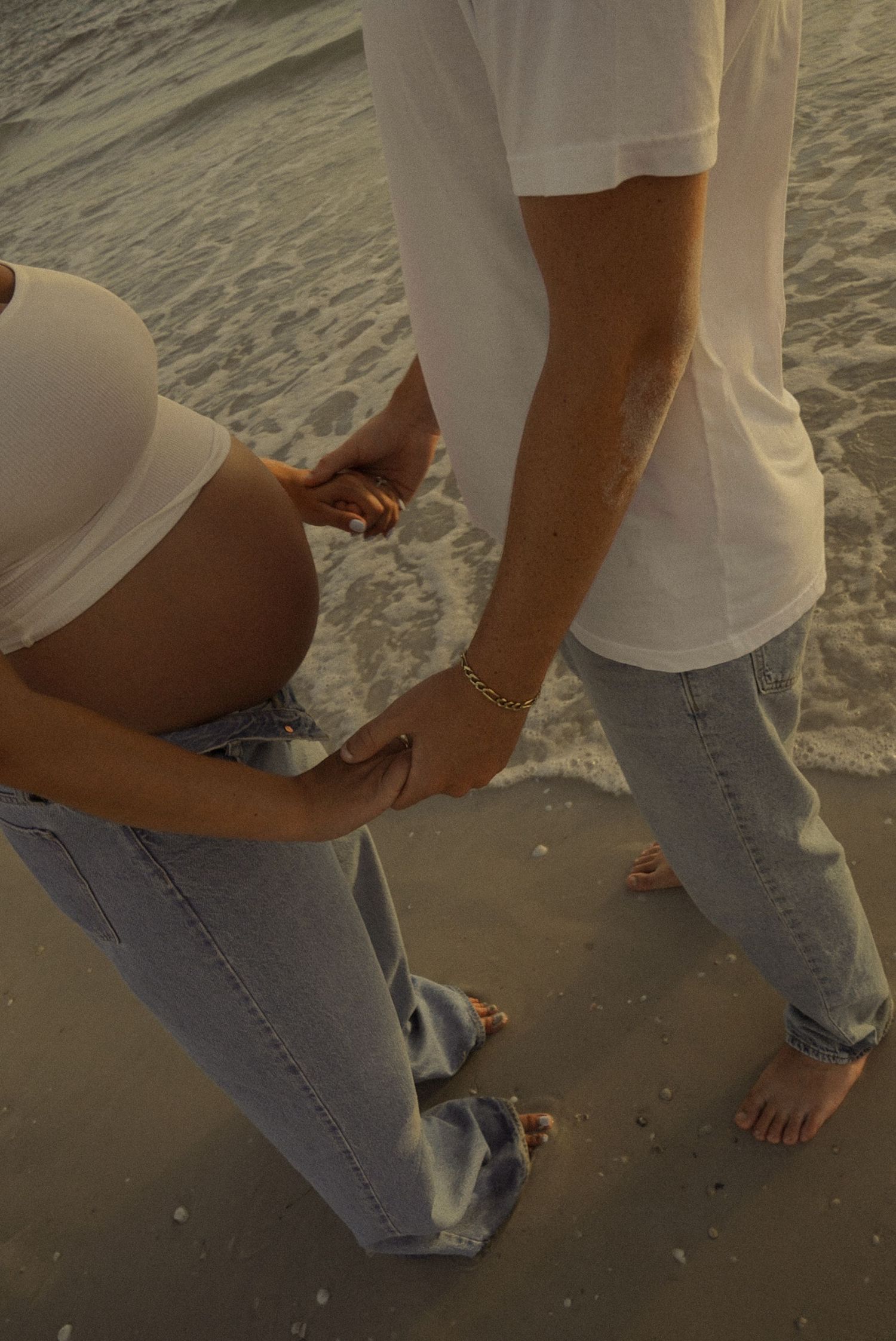 Pregnant woman in white top and jeans holding belly on sandy beach with partner nearby.