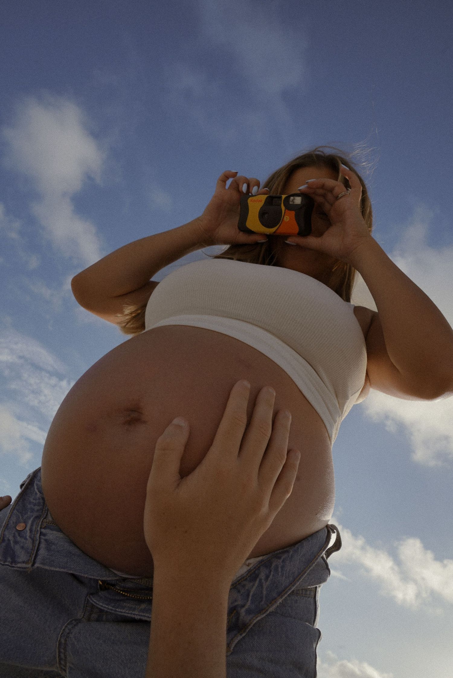 Pregnant woman in white crop top and jeans holds a camera outdoors under a blue sky.