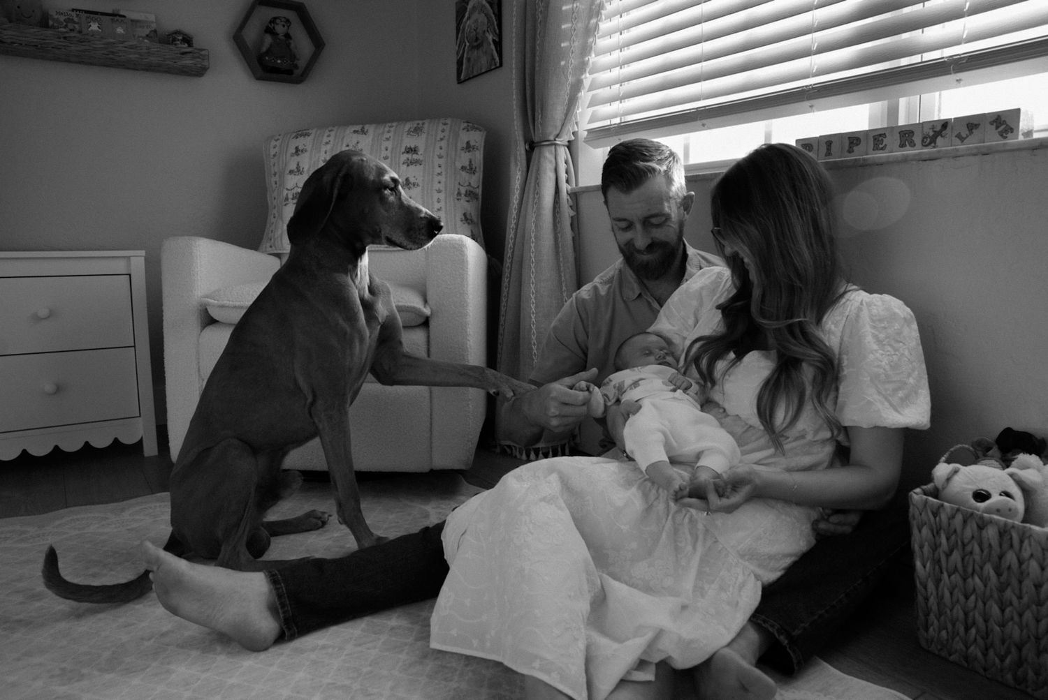 Black and white photo of couple with newborn baby and large dog sitting together in a nursery room.