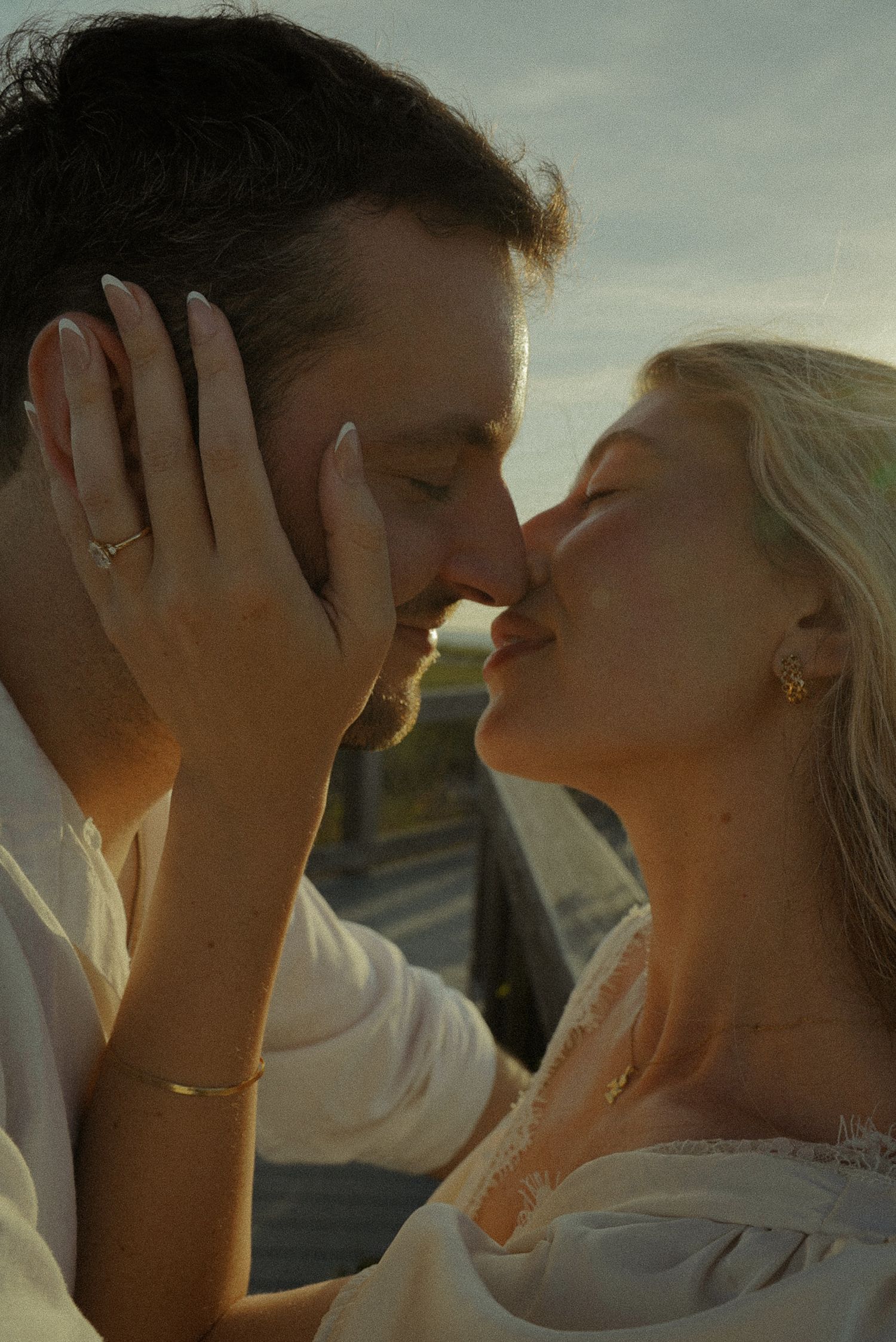 A couple shares a romantic kiss outdoors during golden hour, the woman wearing a delicate bracelet and ring.