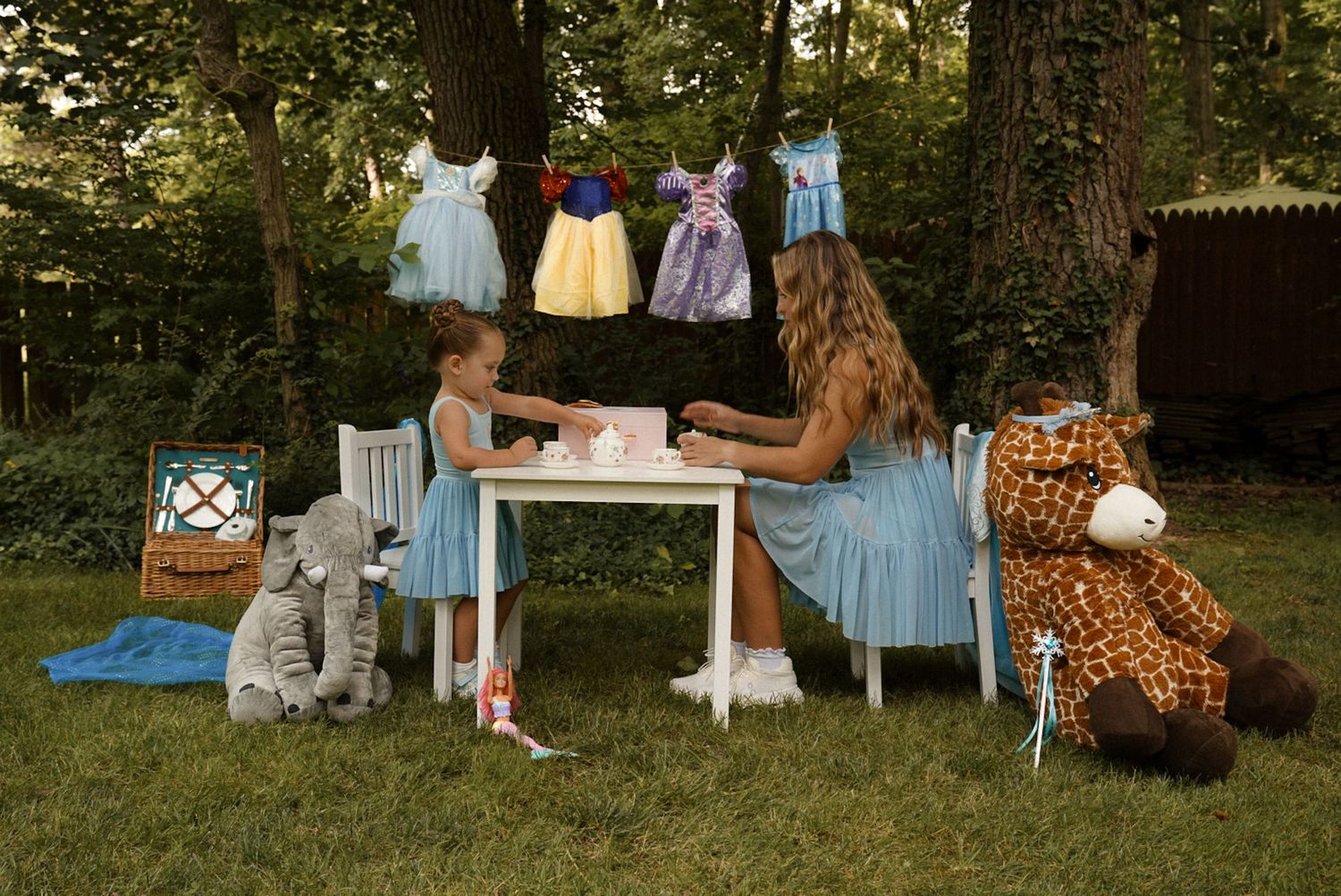Two girls having a tea party outdoors with stuffed animals and princess dresses hanging from a tree.