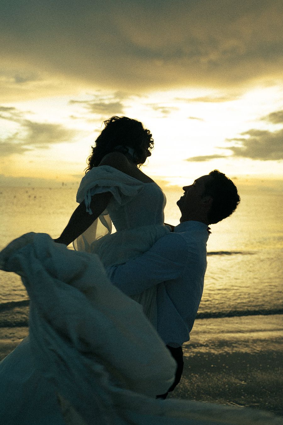 A groom lifts his bride in a flowing white dress on a beach at sunset, silhouetted against a golden sky.