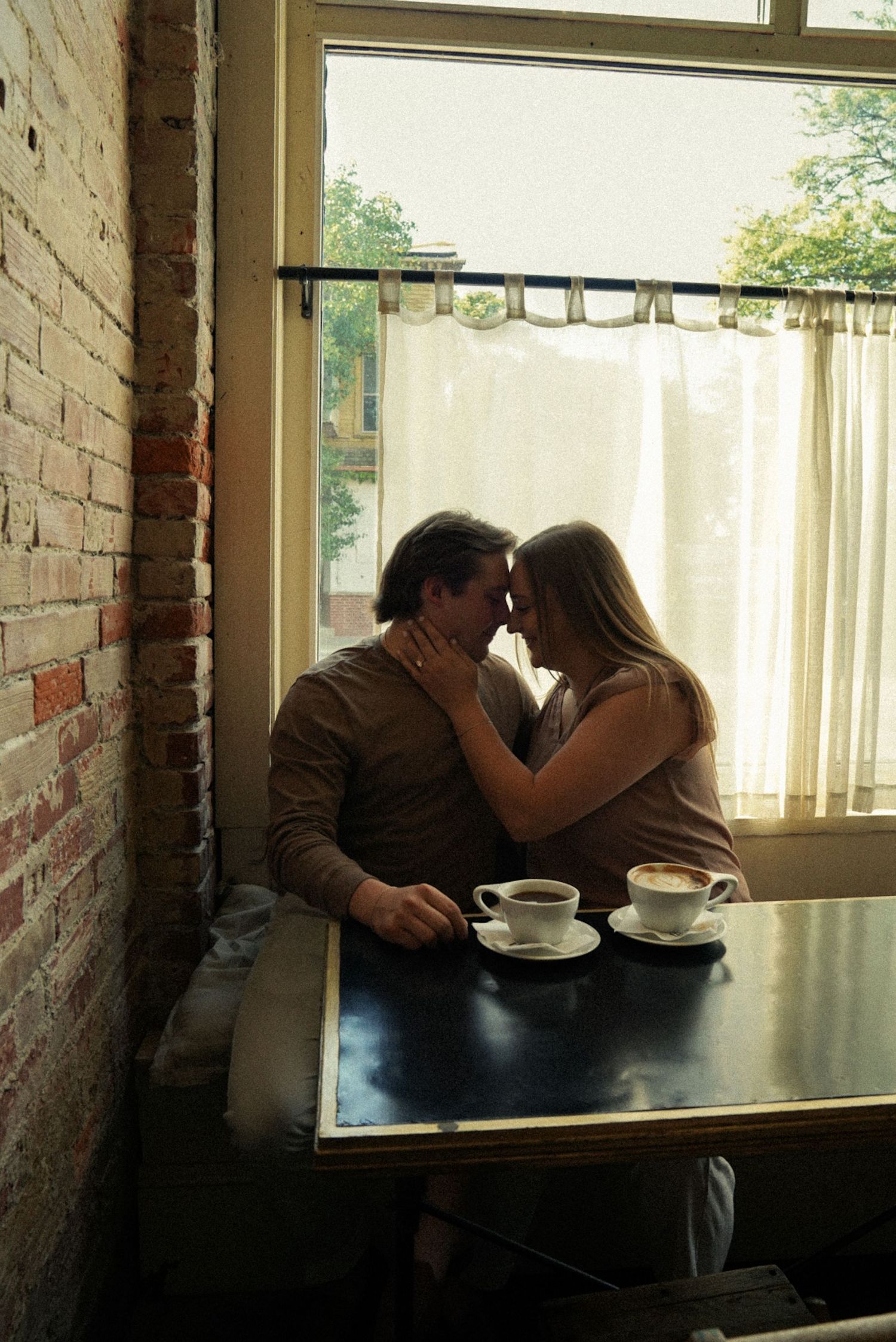 Romantic couple sharing an intimate moment over coffee cups at a cafe table by a curtained window with brick wall.