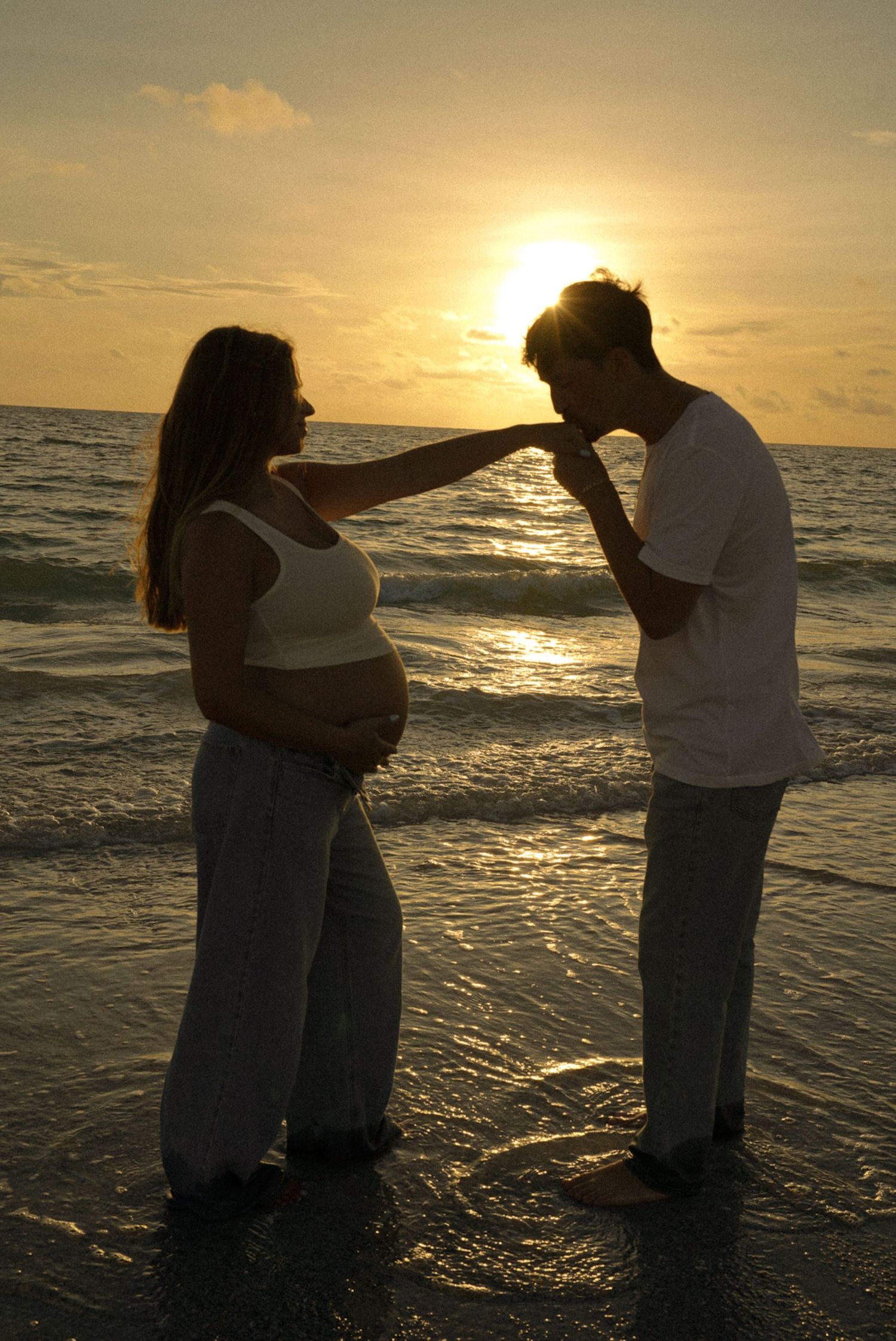 Pregnant woman and man sharing a romantic moment on the beach at sunset, silhouetted against golden sky.