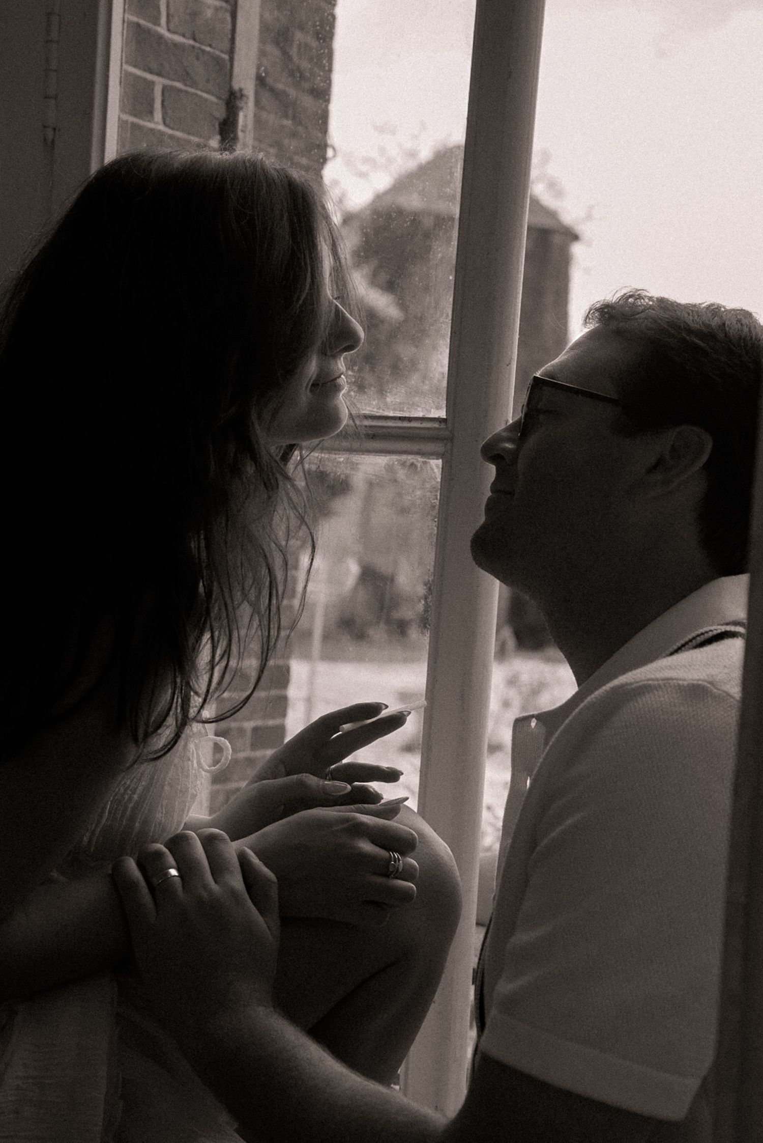 Black and white photo of a couple sharing an intimate moment by a window, gazing at each other tenderly.
