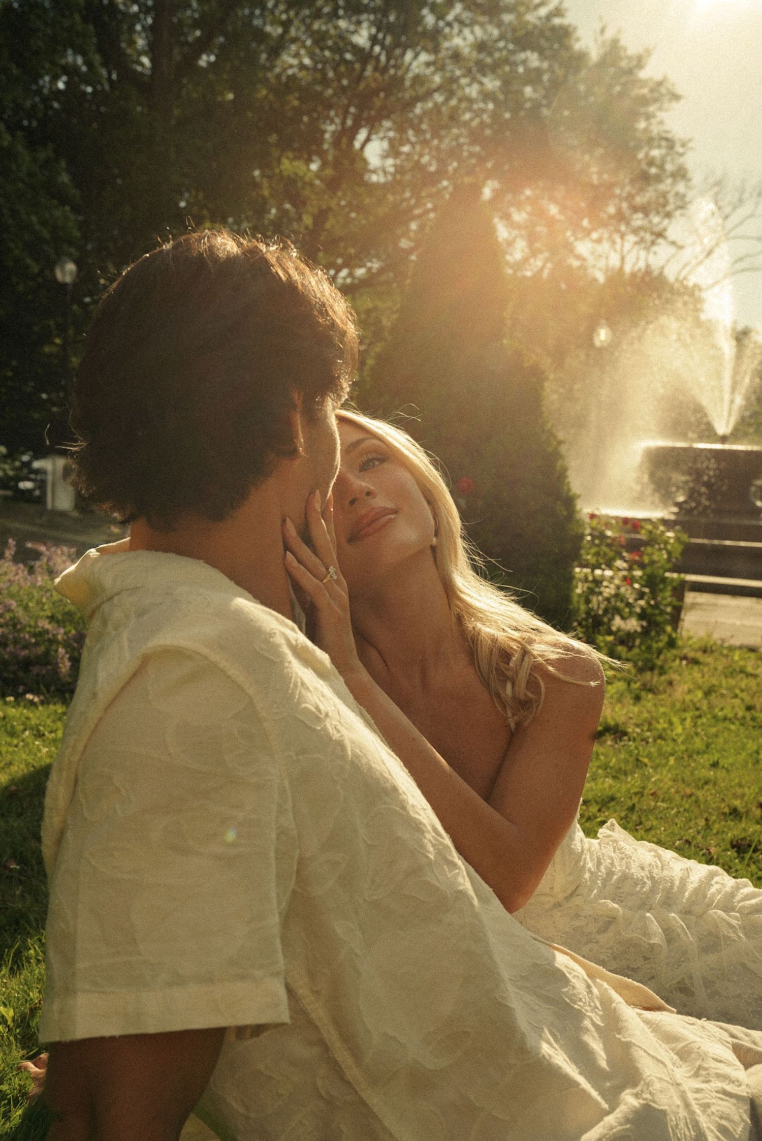 A couple shares a romantic moment outdoors, bathed in golden sunlight near a fountain.