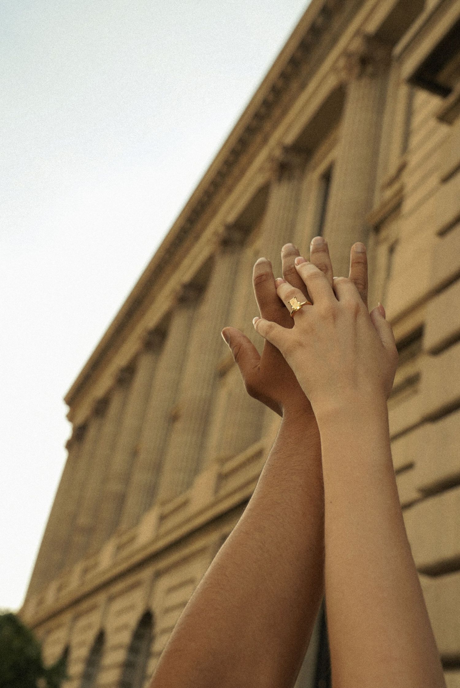 Two hands raised together near a classical building, one adorned with gold rings, against a soft sky.