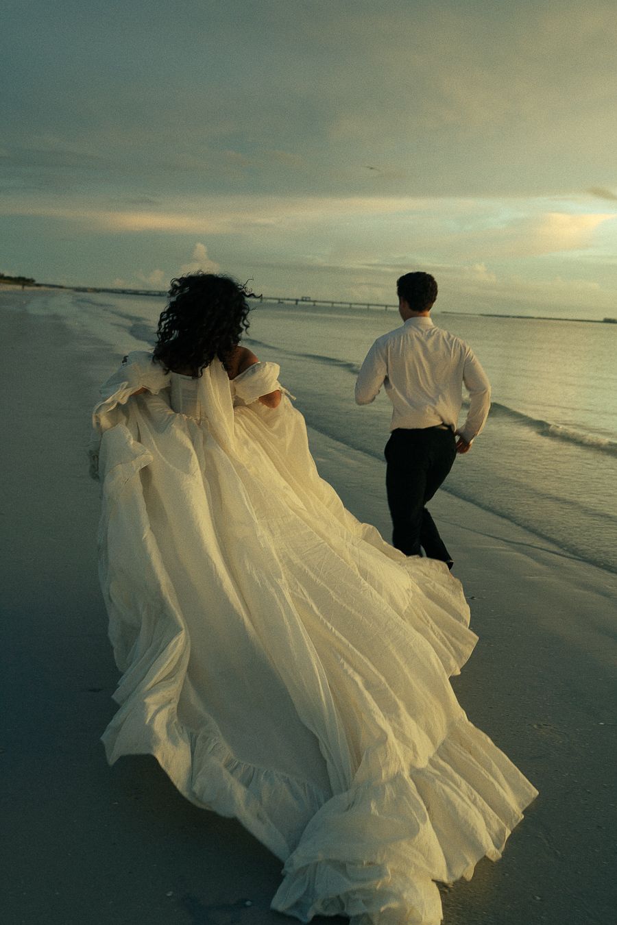 Bride in flowing white gown and groom in black pants run along a sandy beach at sunset.
