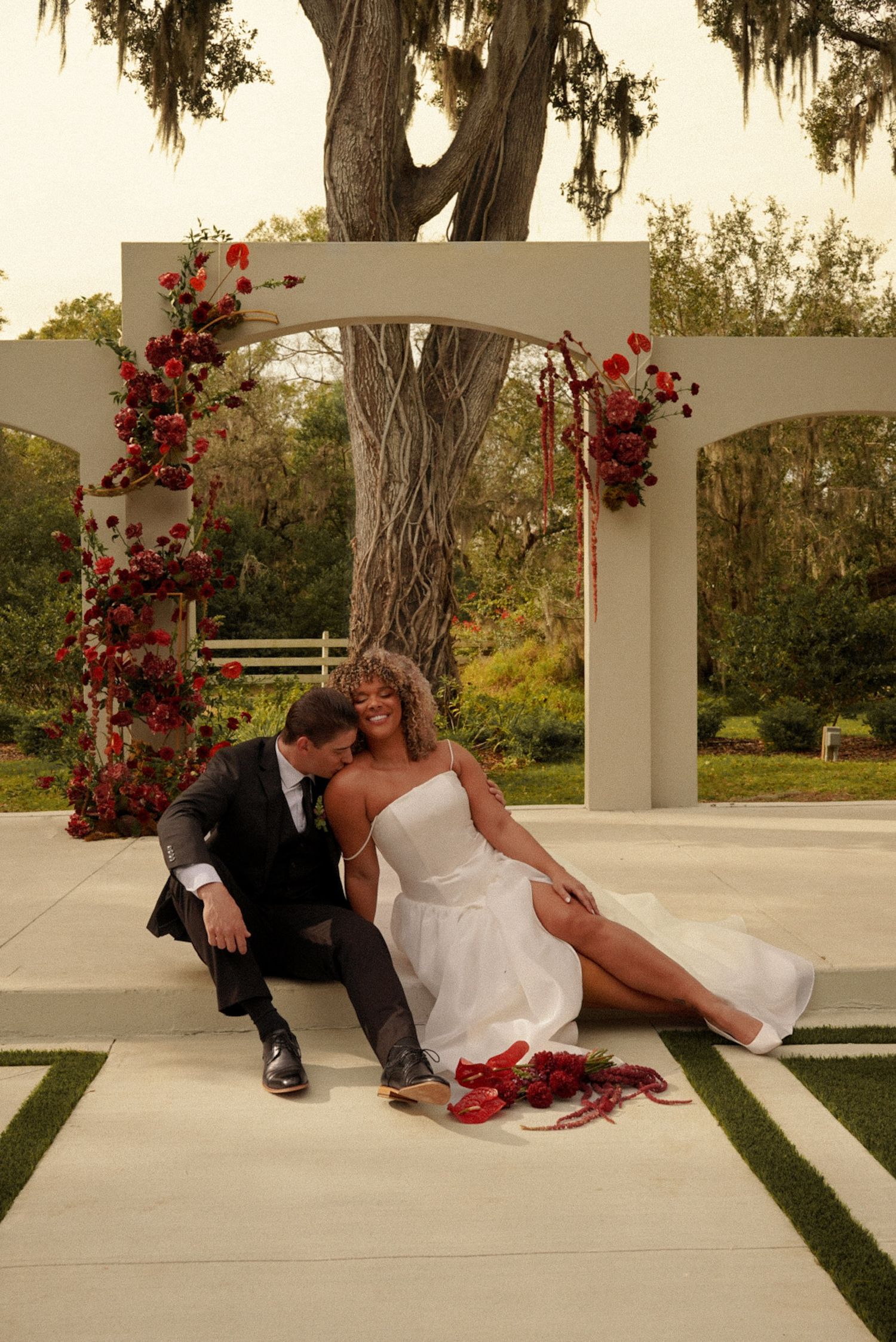 Bride and groom sit together under a floral arch adorned with red roses at an outdoor wedding ceremony.