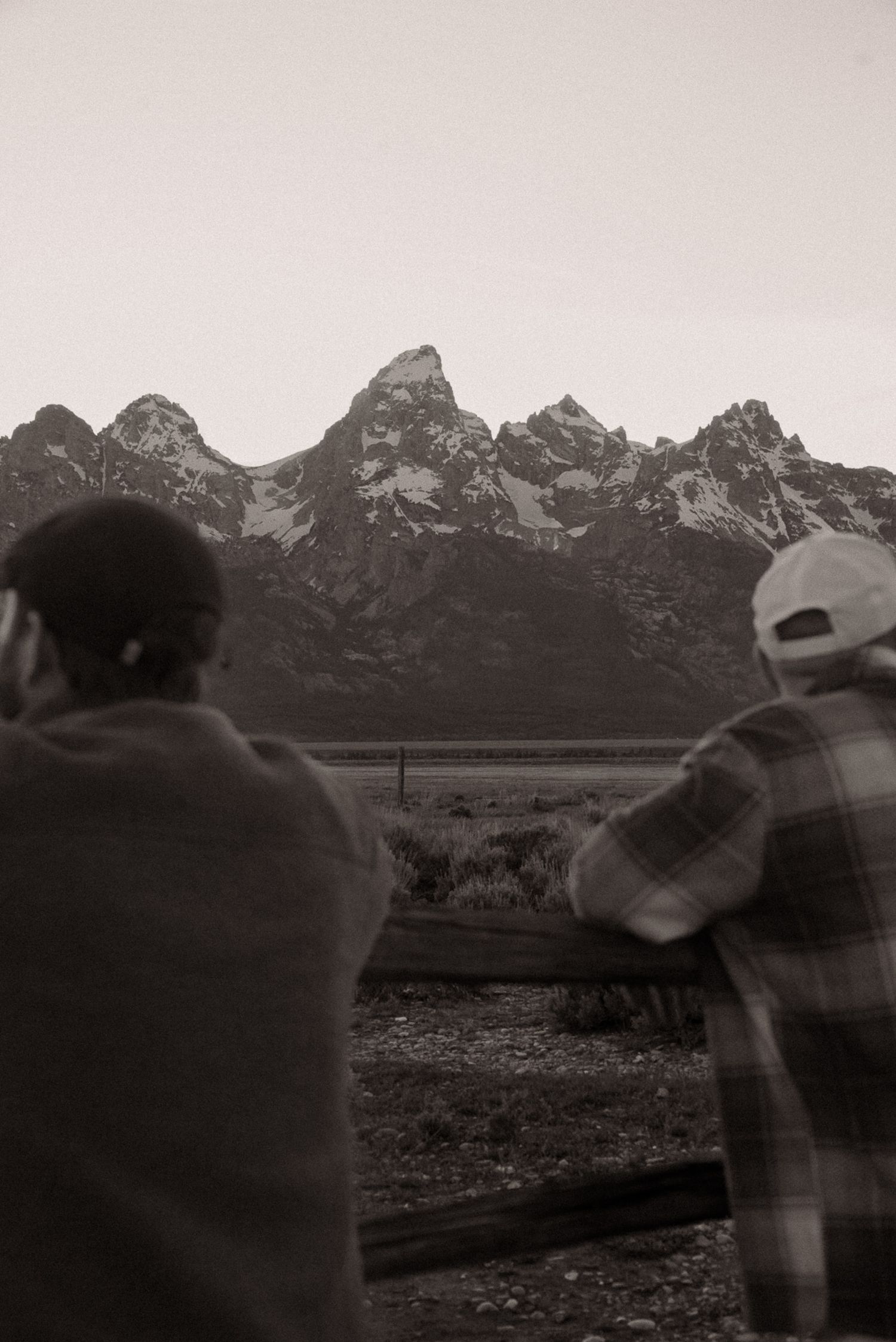Two people wrapped in blankets gaze at snow-capped Teton mountain range in black and white photo.