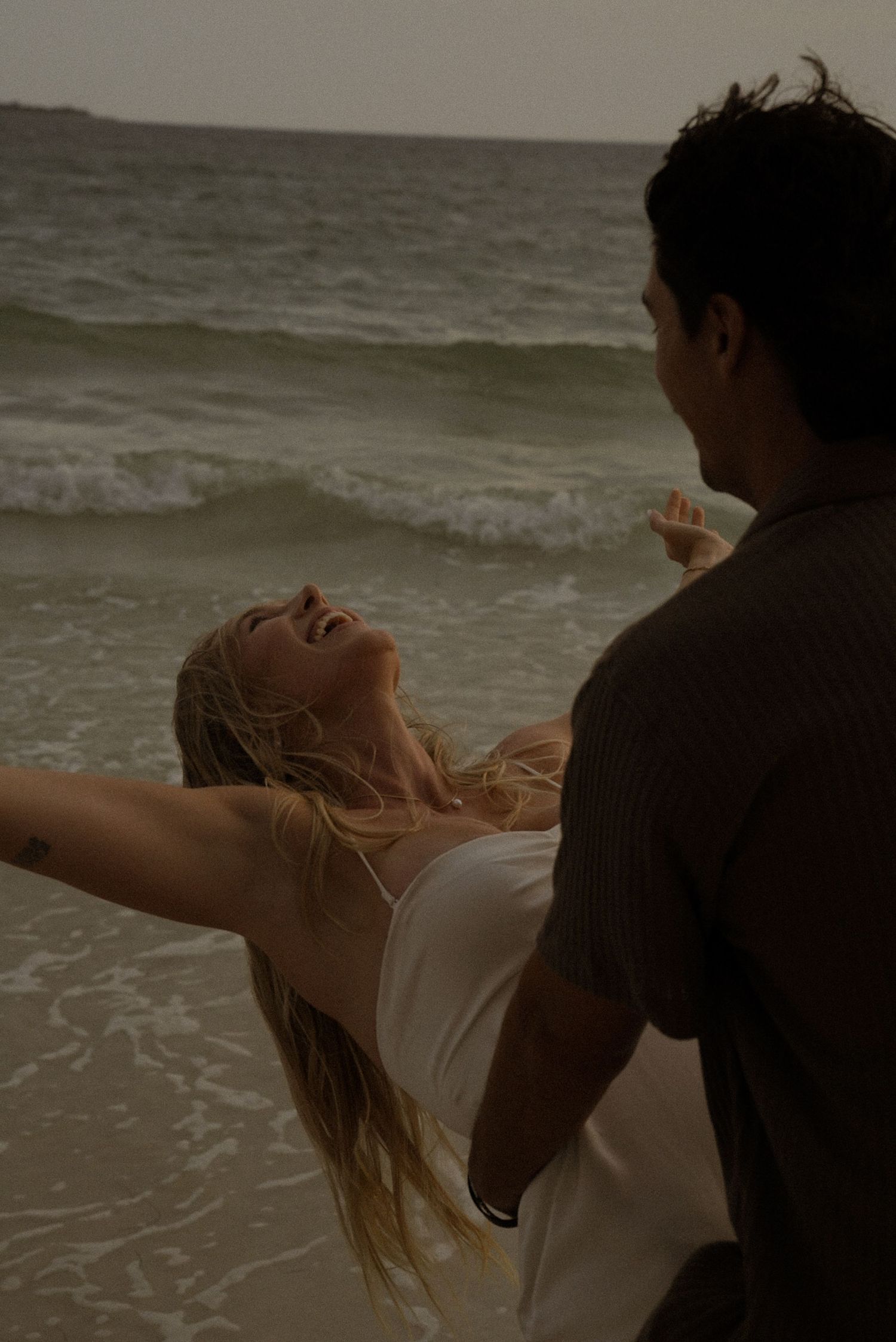 Couple dancing romantically on a beach at dusk, woman in white dress laughing with arms outstretched near ocean waves.