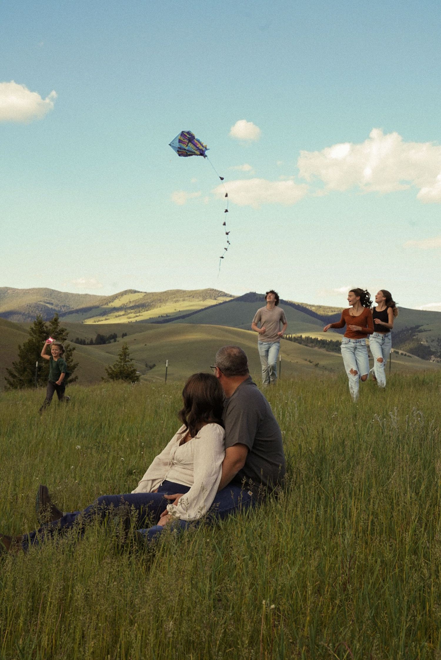 Family flying a colorful kite on a green hillside, couple sitting in grass while children run in scenic mountain landscape.