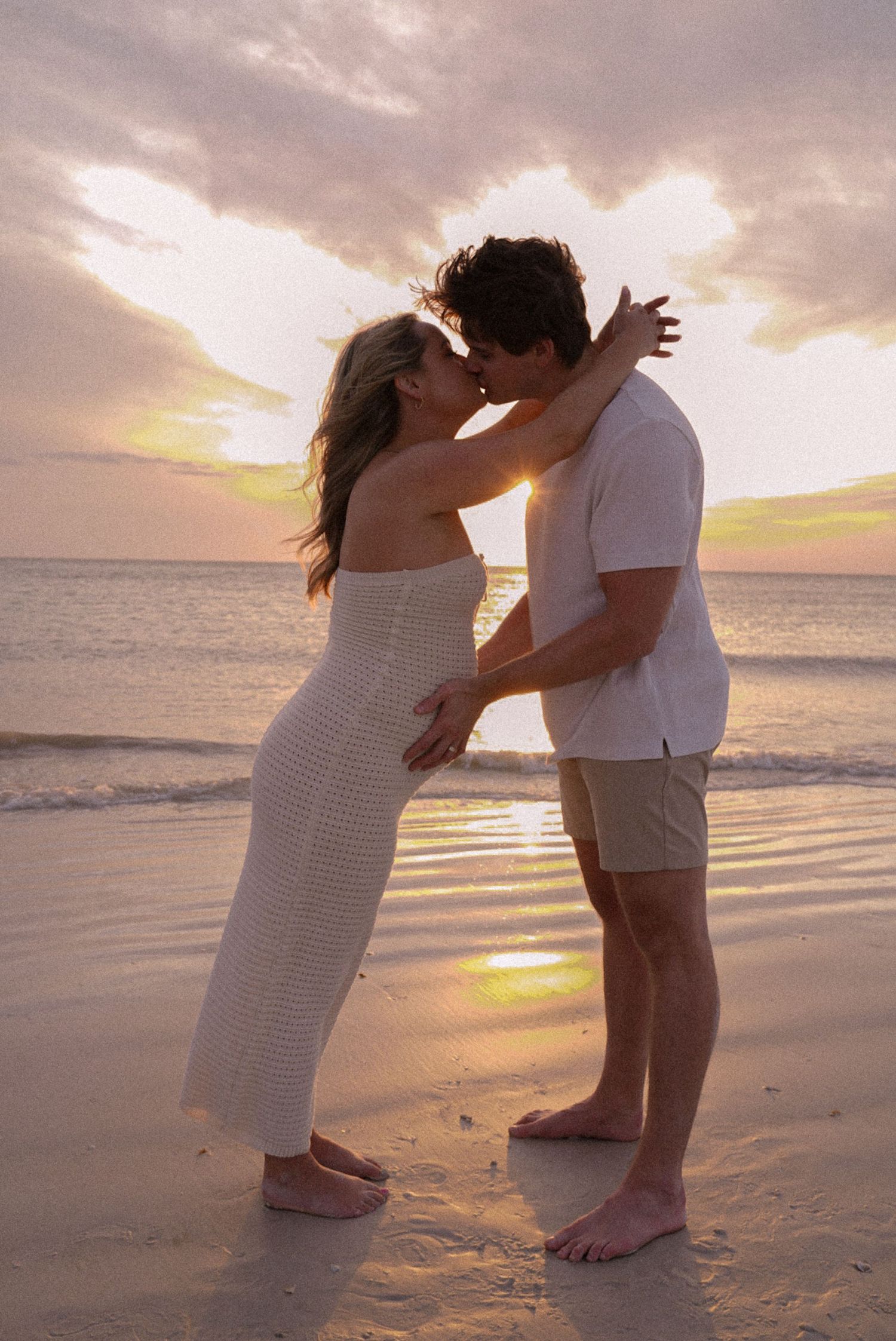 A couple shares a romantic kiss on a sandy beach at sunset, with golden light reflecting off the calm ocean waves.